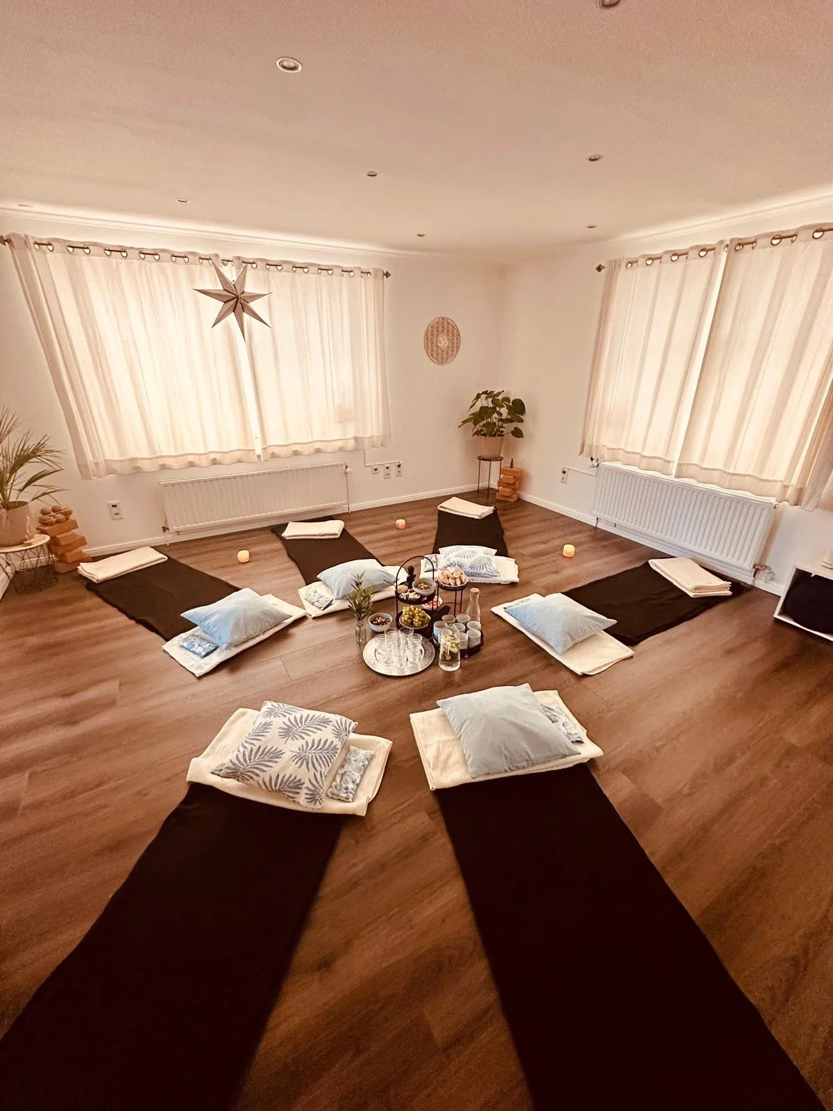 Das Bild zeigt einen Raum mit einer Bodenmassage- oder Meditationsrunde, bei der Matten, Kissen und Decken in einem kreisförmigen Arrangement liegen. Es gibt Getränke, Kerzen und kleine Dekorationen auf einem Tablett in der Mitte des Kreises, und der Raum ist hell erleuchtet mit großen Fenstern und weißen Vorhängen.