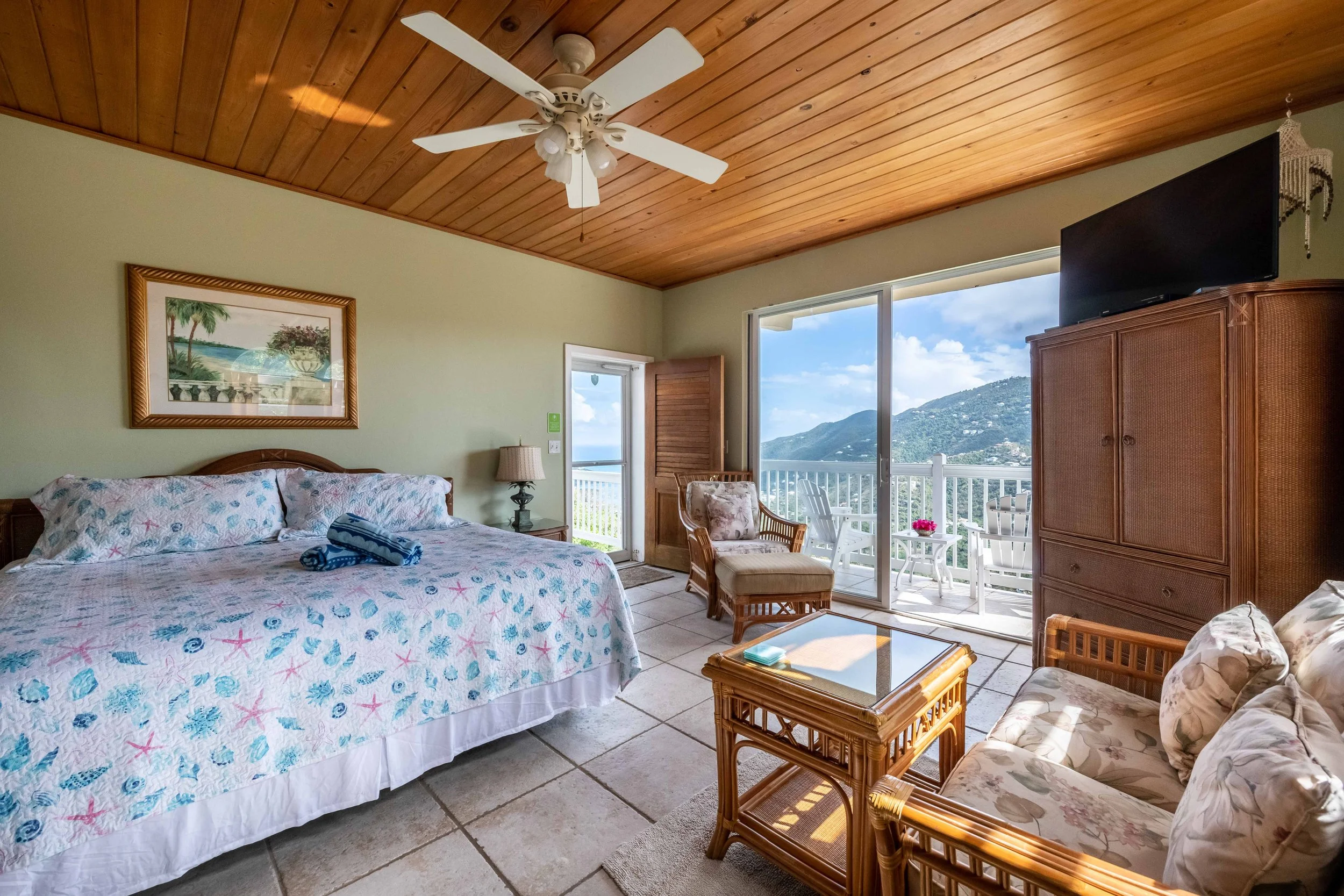 Coastal bedroom with wooden ceiling, ceiling fan, framed landscape picture, sliding glass door leading to balcony with mountain view, wicker furniture, and tropical decor.