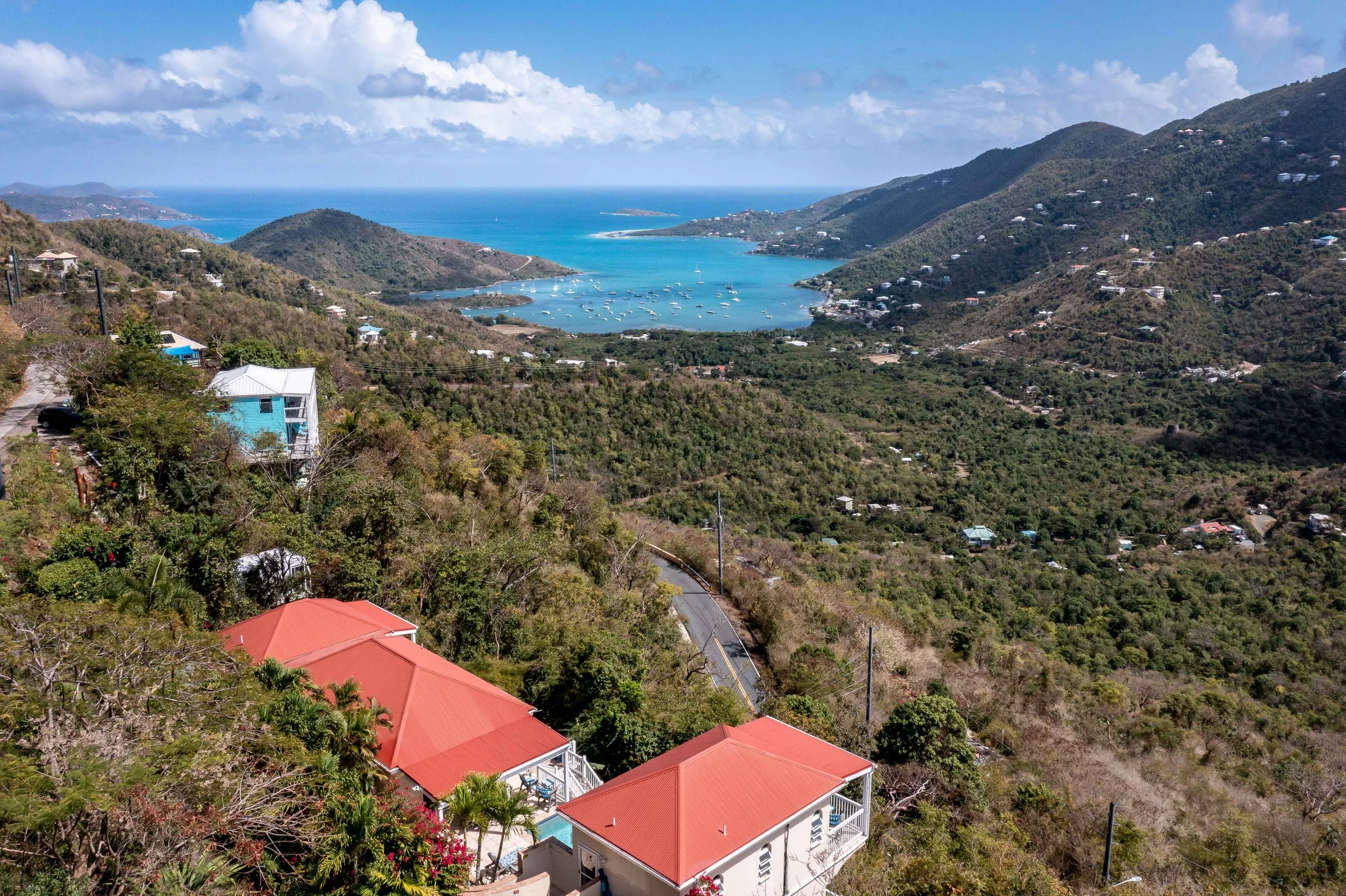 Scenic view of a coastal bay surrounded by green hills with houses, boats in the water, and a blue sky with clouds.