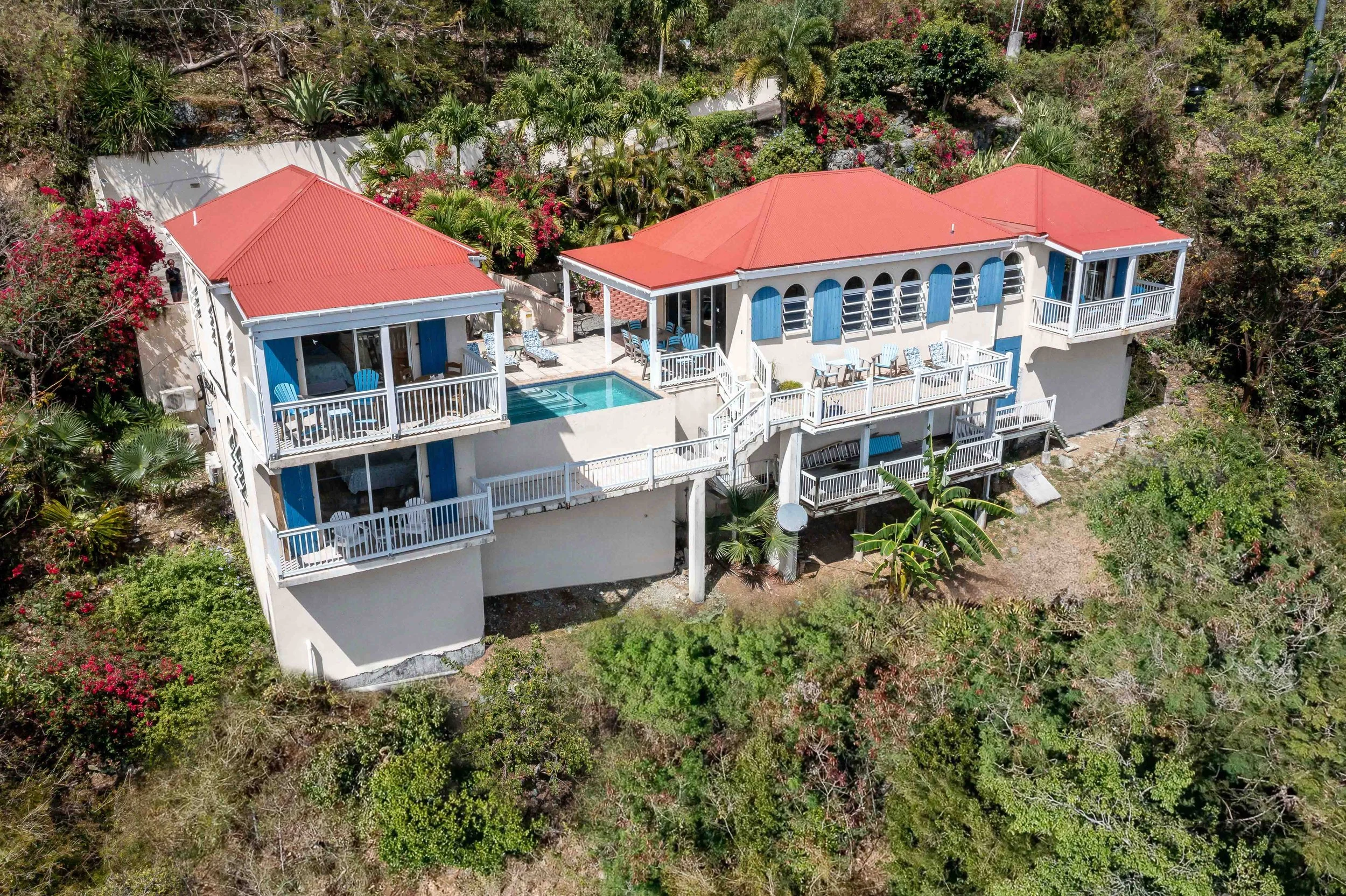 Aerial view of a house with red tiled roof, white walls, and multiple balconies surrounded by lush greenery and colorful flowering plants.