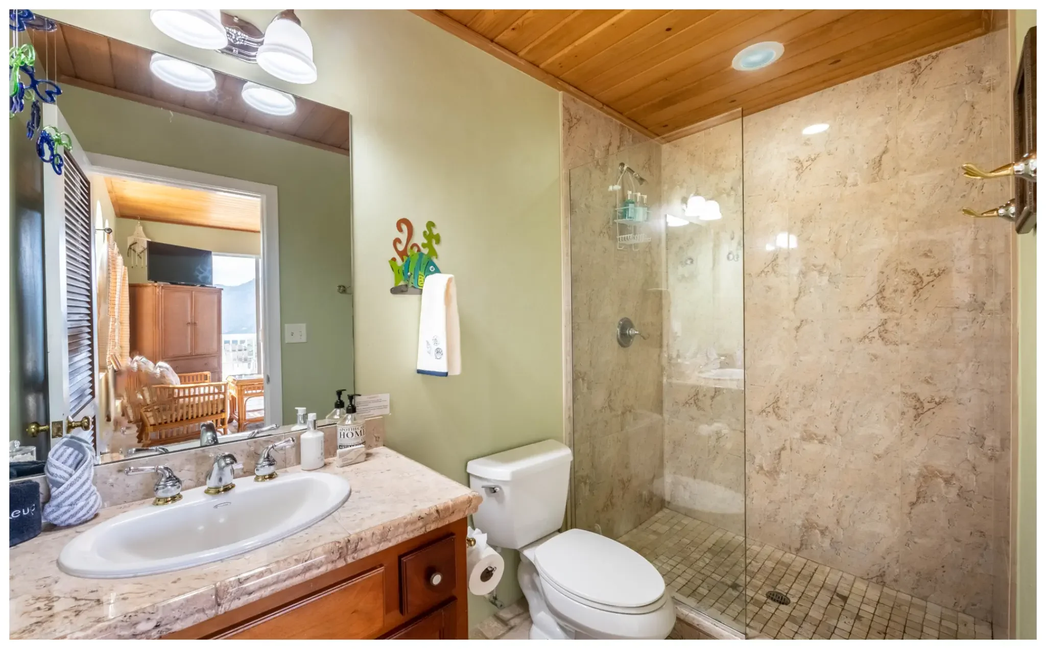 A bathroom with a marble countertop, a white sink, and a wooden vanity cabinet. There are bottles and toiletries on the counter, a towel hanging on a hook, and a toilet next to the shower area. The shower has a glass door, beige marble tiles, and a small stone floor. A wooden ceiling and a mirror above the sink reflect part of the room and a doorway leading to a sunlit room with wooden furniture and a TV.