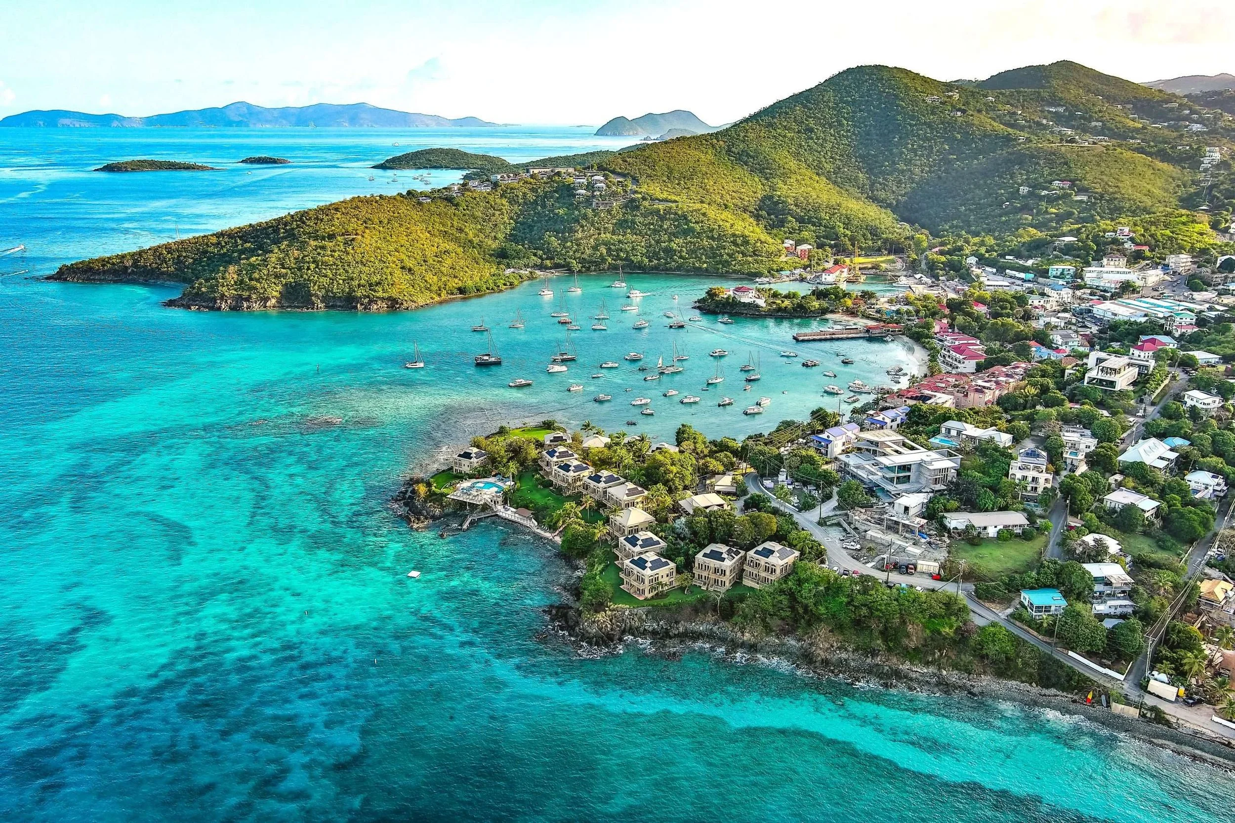 Aerial view of a coastal town with boats docked in a turquoise bay, surrounded by green hills and residential buildings, with mountains in the background.