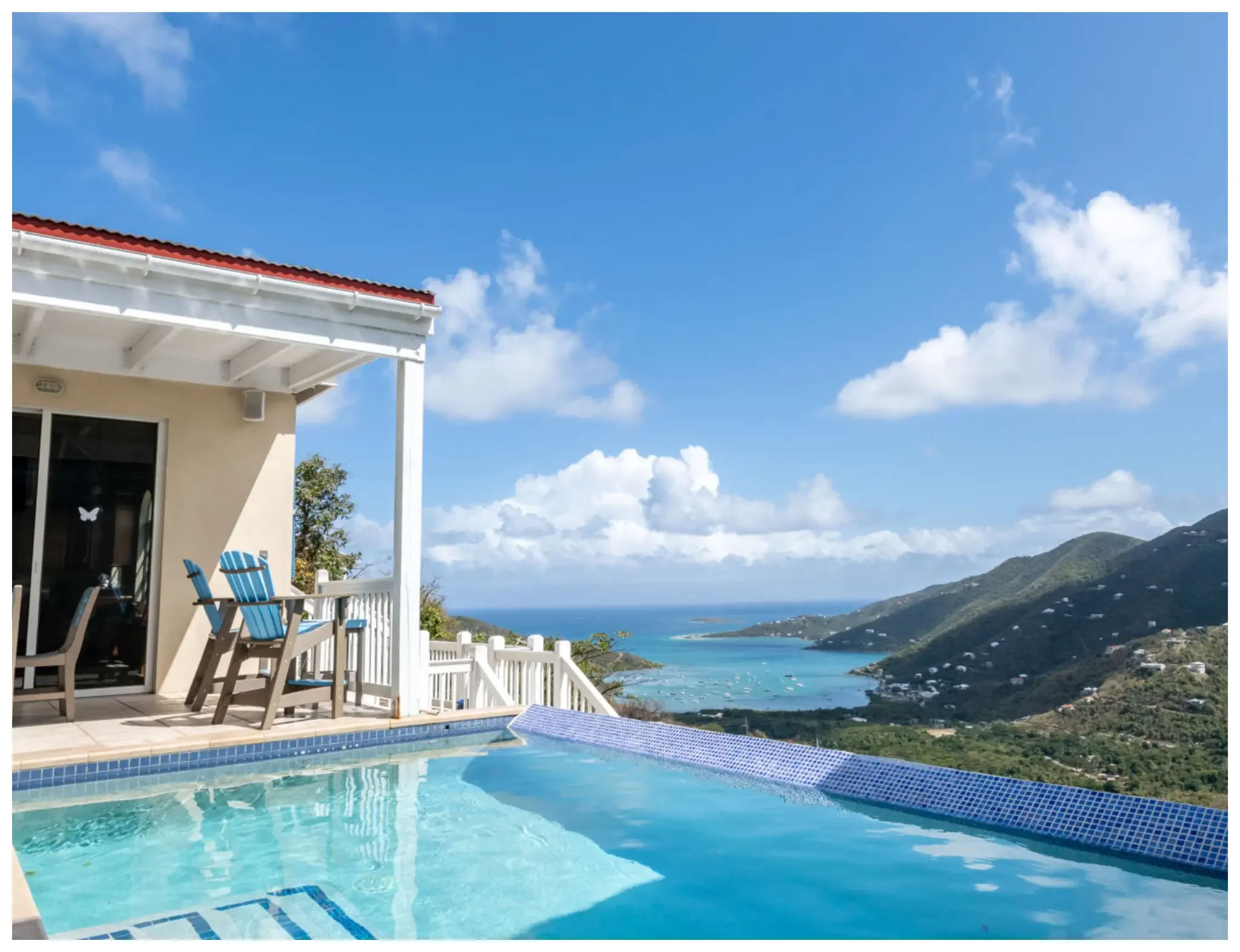 View of a house with a patio, a swimming pool, and a scenic ocean view with mountains under a blue sky with some clouds.