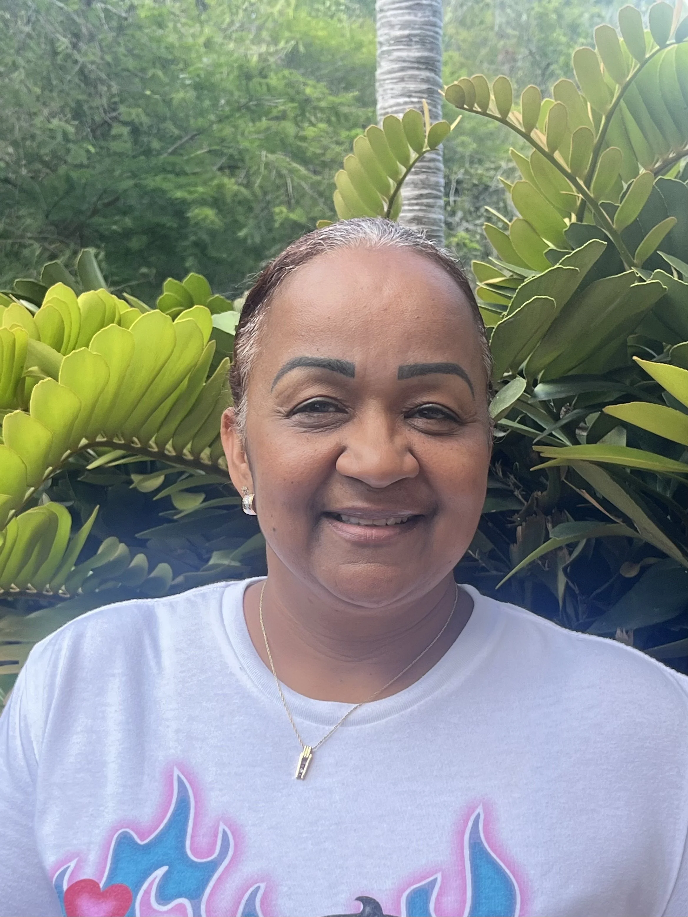 A woman with gray hair and earrings smiling in front of lush green plants and trees.