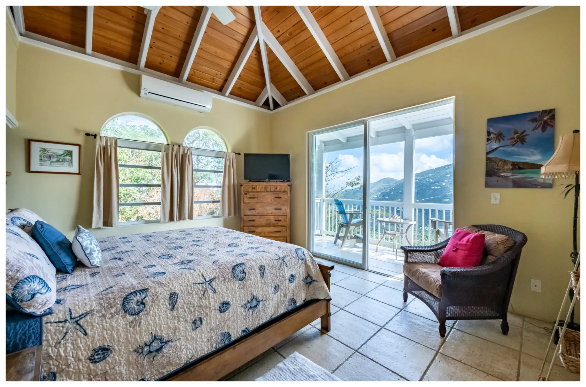 A bedroom with a vaulted wooden ceiling, yellow walls, a bed with ocean-themed quilted bedding, a wicker chair with a red cushion, and a sliding glass door leading to a balcony with mountain views.