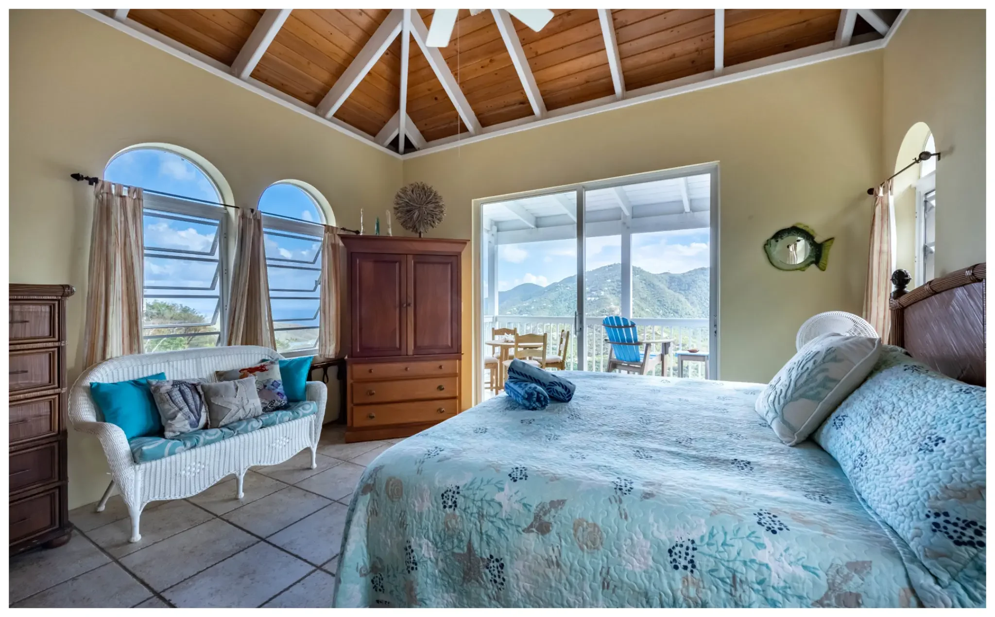 A bright bedroom with a vaulted wood ceiling, pale yellow walls, and large stained-glass windows showing a mountain landscape. The room has a white wicker loveseat with colorful pillows, a wooden dresser, and a bed with a patterned quilt. There are small tables and chairs on the balcony outside, visible through sliding glass doors.