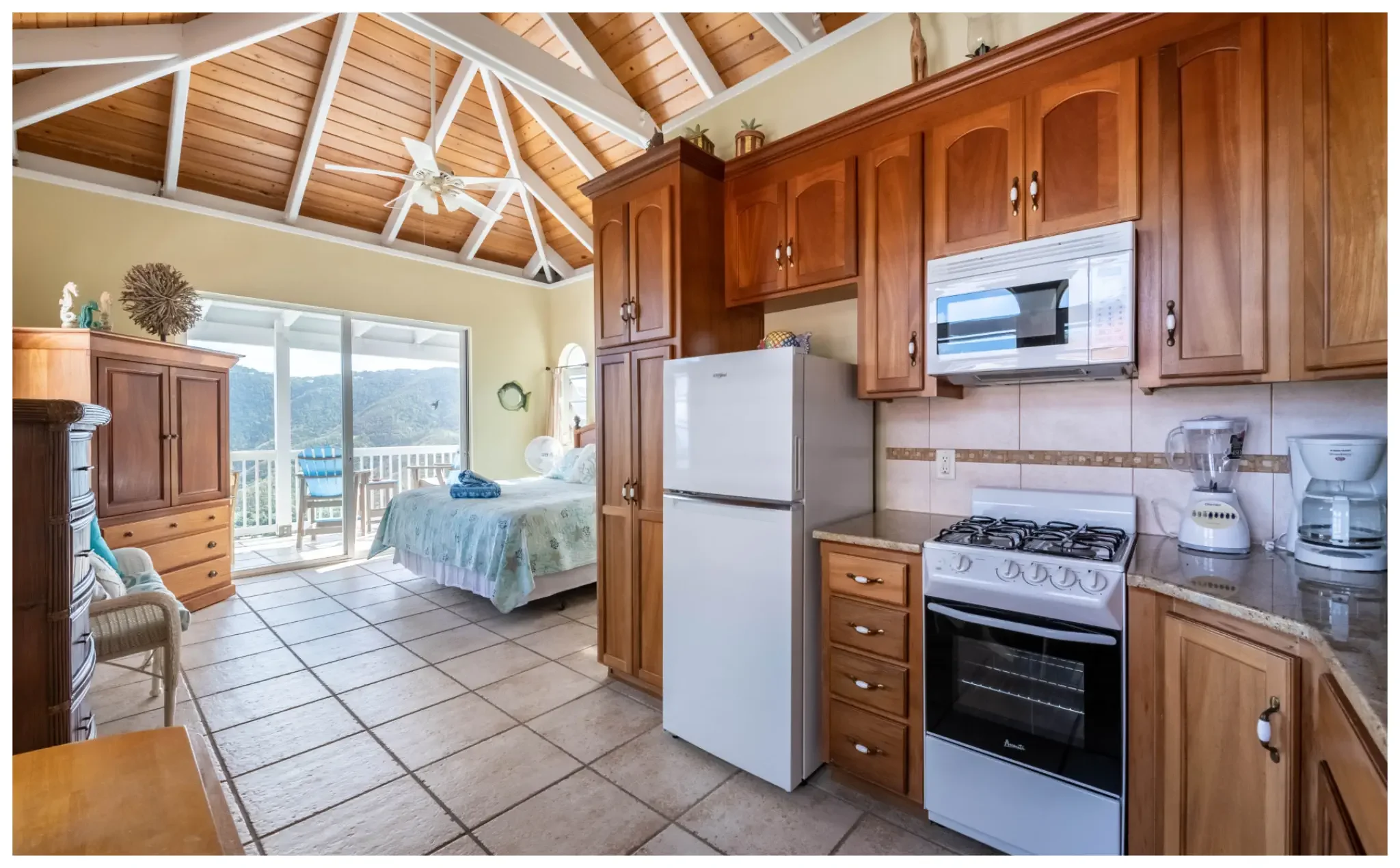 Open-concept kitchen and bedroom area with wood cabinets, white appliances, tiled floor, and a sliding glass door leading to a balcony with mountain view.