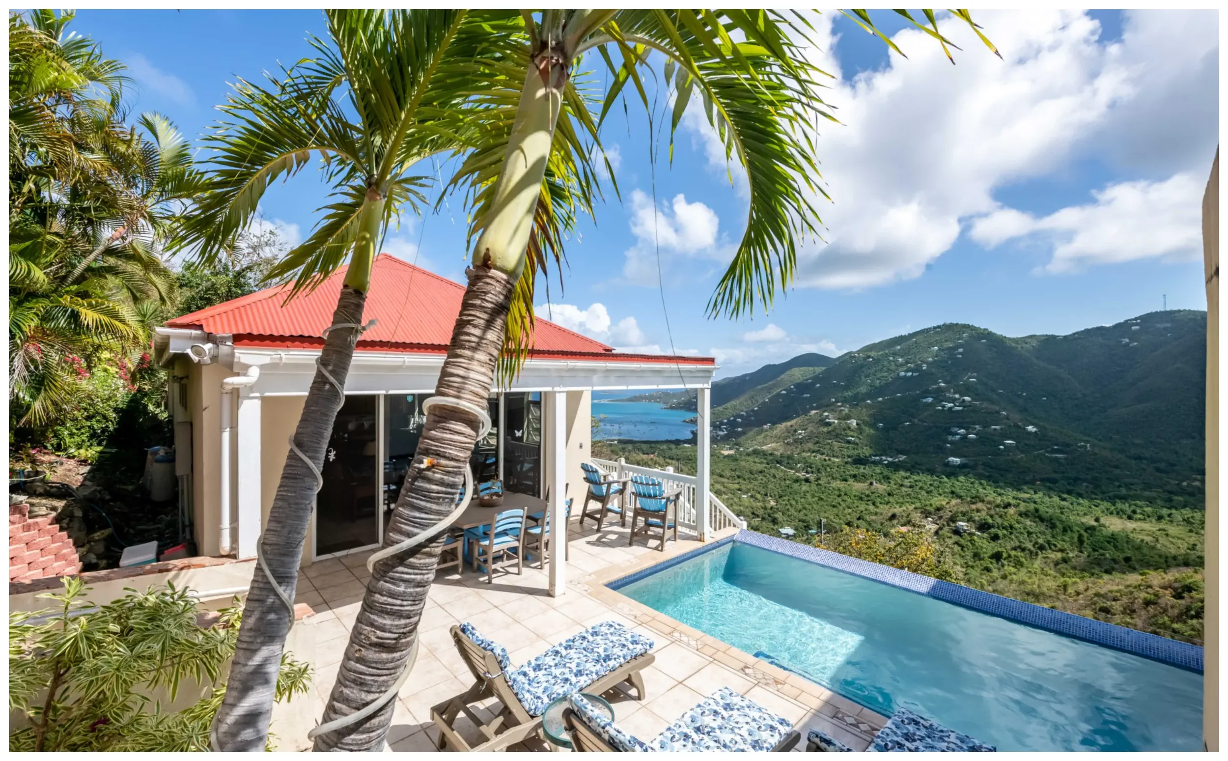 View of a backyard with a small swimming pool, outdoor dining table with chairs, lounge chairs with blue and white cushions, surrounded by lush green vegetation and palm trees, with a house that has a red roof and mountains in the background under a partly cloudy sky.