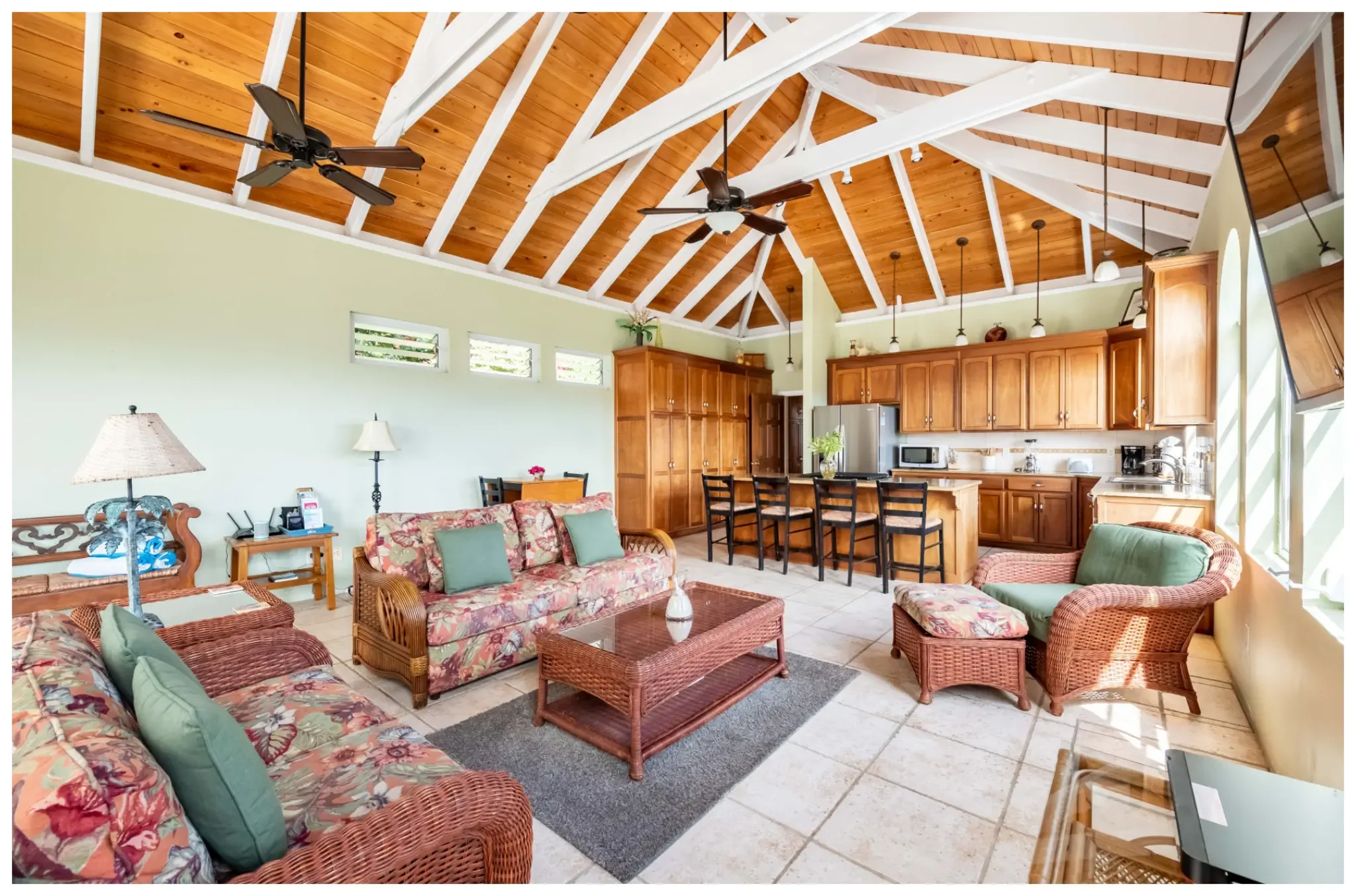 Living room with tropical-style furniture and an open kitchen with wooden cabinets, tiled floor, ceiling fans, and a vaulted wooden ceiling with white beams.