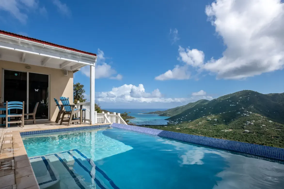 A balcony with a swimming pool overlooking green hillside and ocean with clouds in the sky.