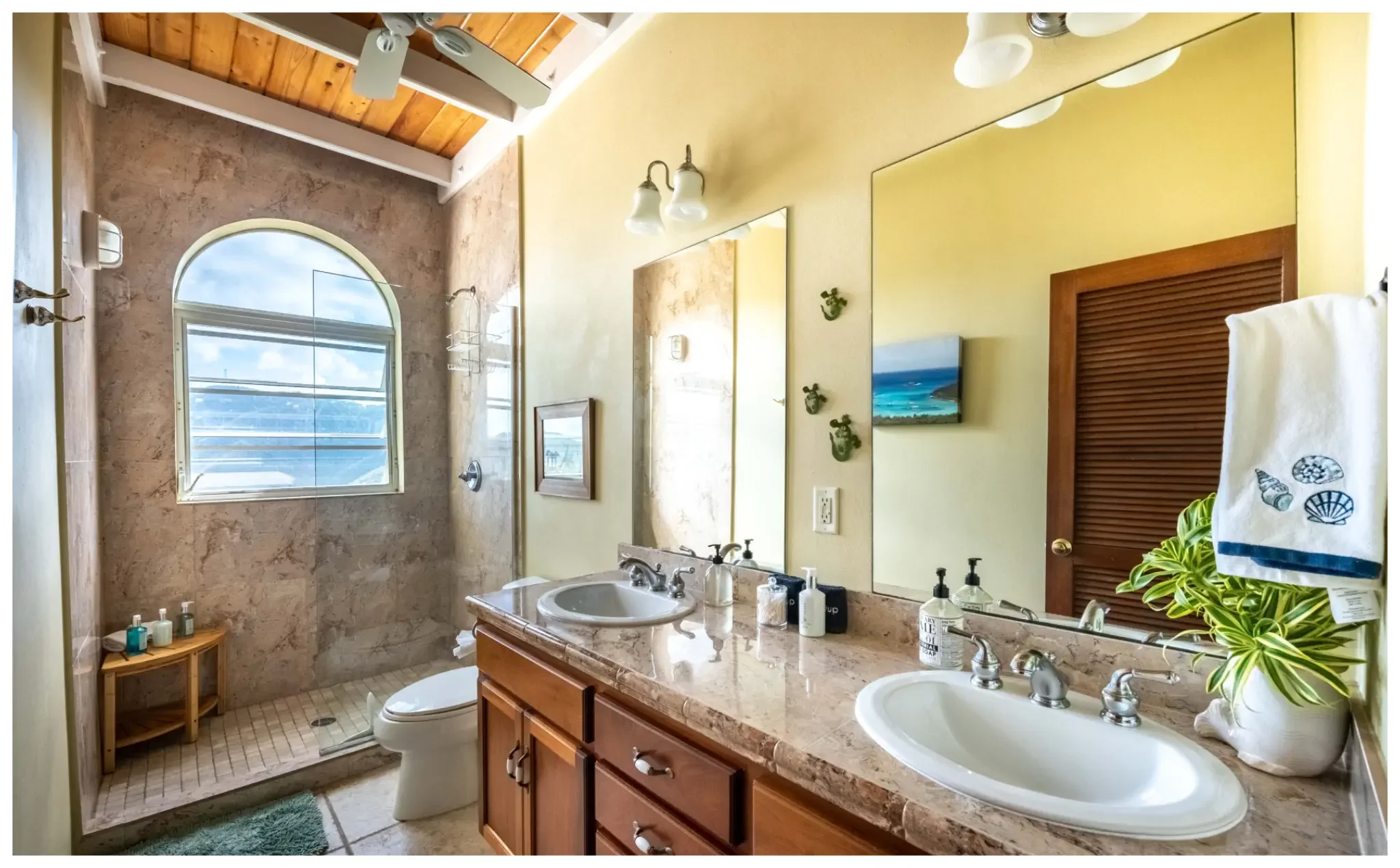 A bathroom with a double sink vanity, beige marble countertops, large mirror, and a window with a view of the sky and clouds. There is a wooden door with louvered panels, a small shower area with a window, and decorative seashells and plants on the counters and towels.
