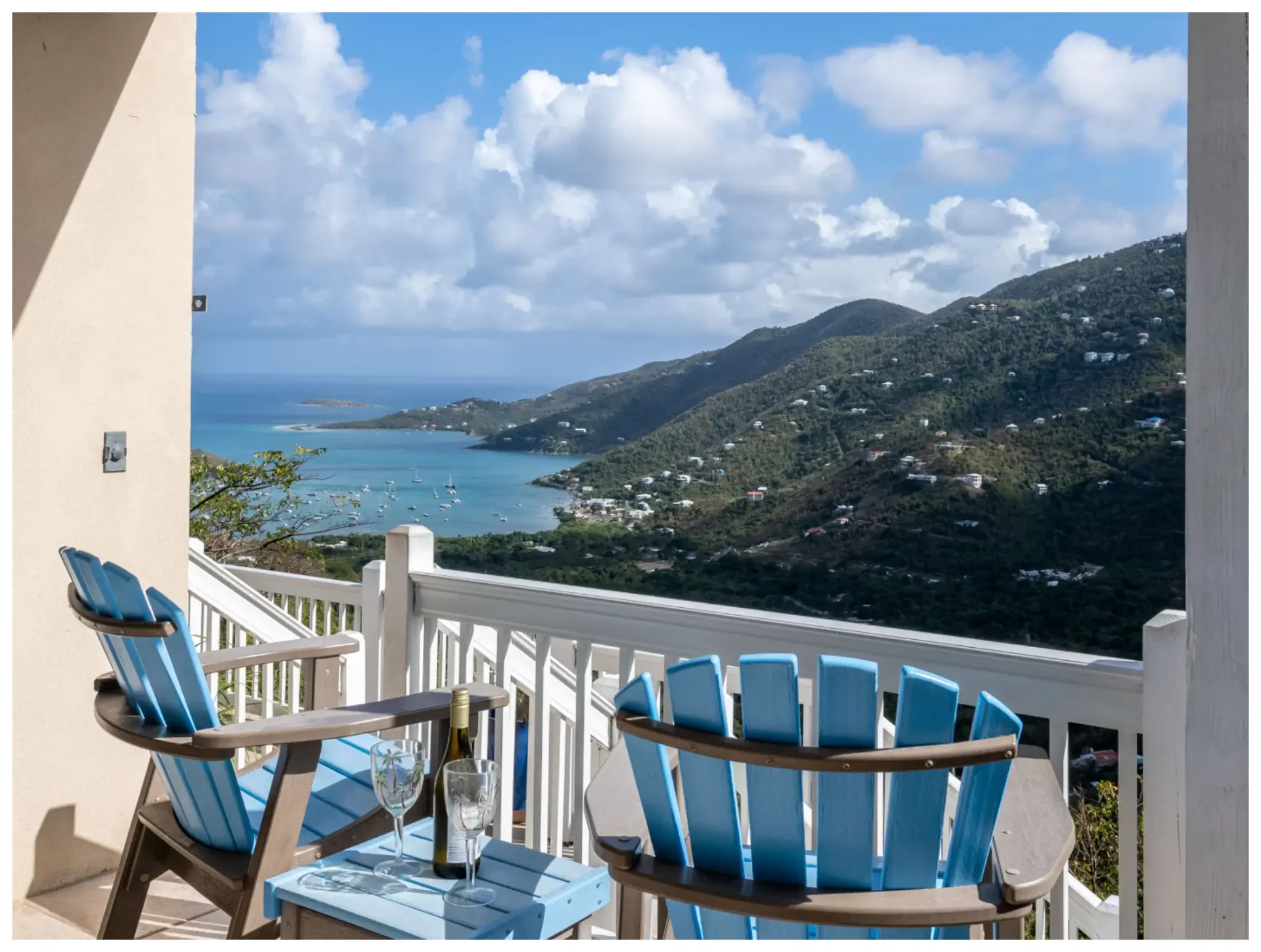Balcony patio with blue Adirondack chairs, a small table with wine and glasses, overlooking a scenic view of the ocean, hills, and cloudy sky.