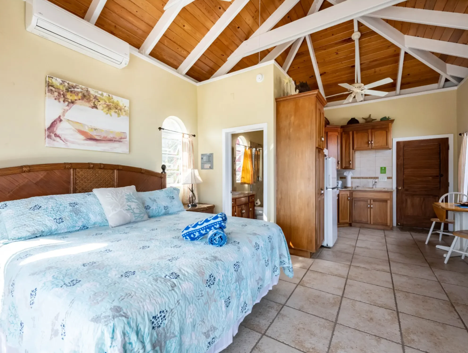 Bedroom with a wooden beamed ceiling, a bed with a white and blue quilt, a bedside lamp, a window with curtains, and a kitchen area in the background.