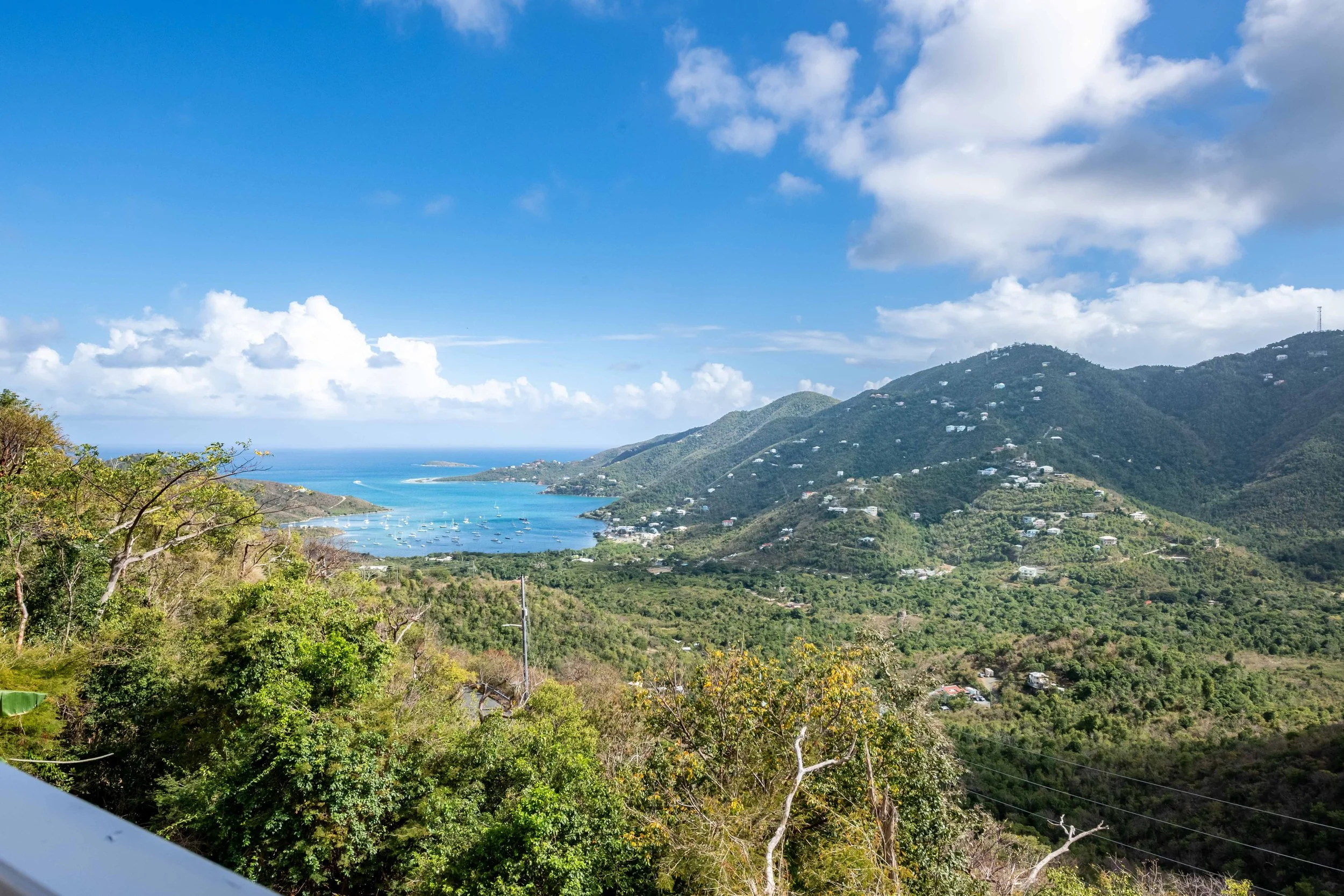 Scenic view of a bay surrounded by green hills with scattered houses, sailboats on the water, and a partly cloudy blue sky.