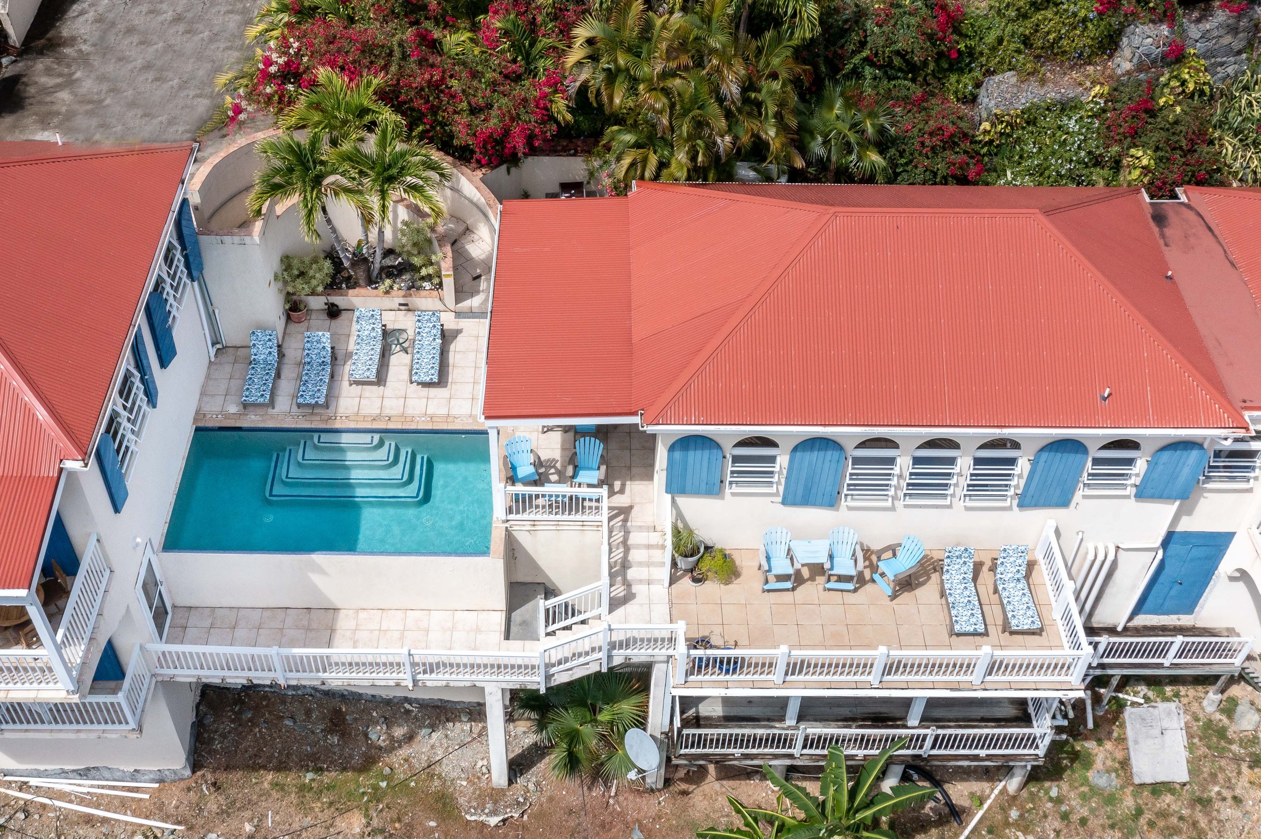 Aerial view of a backyard with a red-tiled roof building on the right, a pool with steps, and outdoor furniture including tables and chairs. The yard is surrounded by lush greenery and flowering plants.