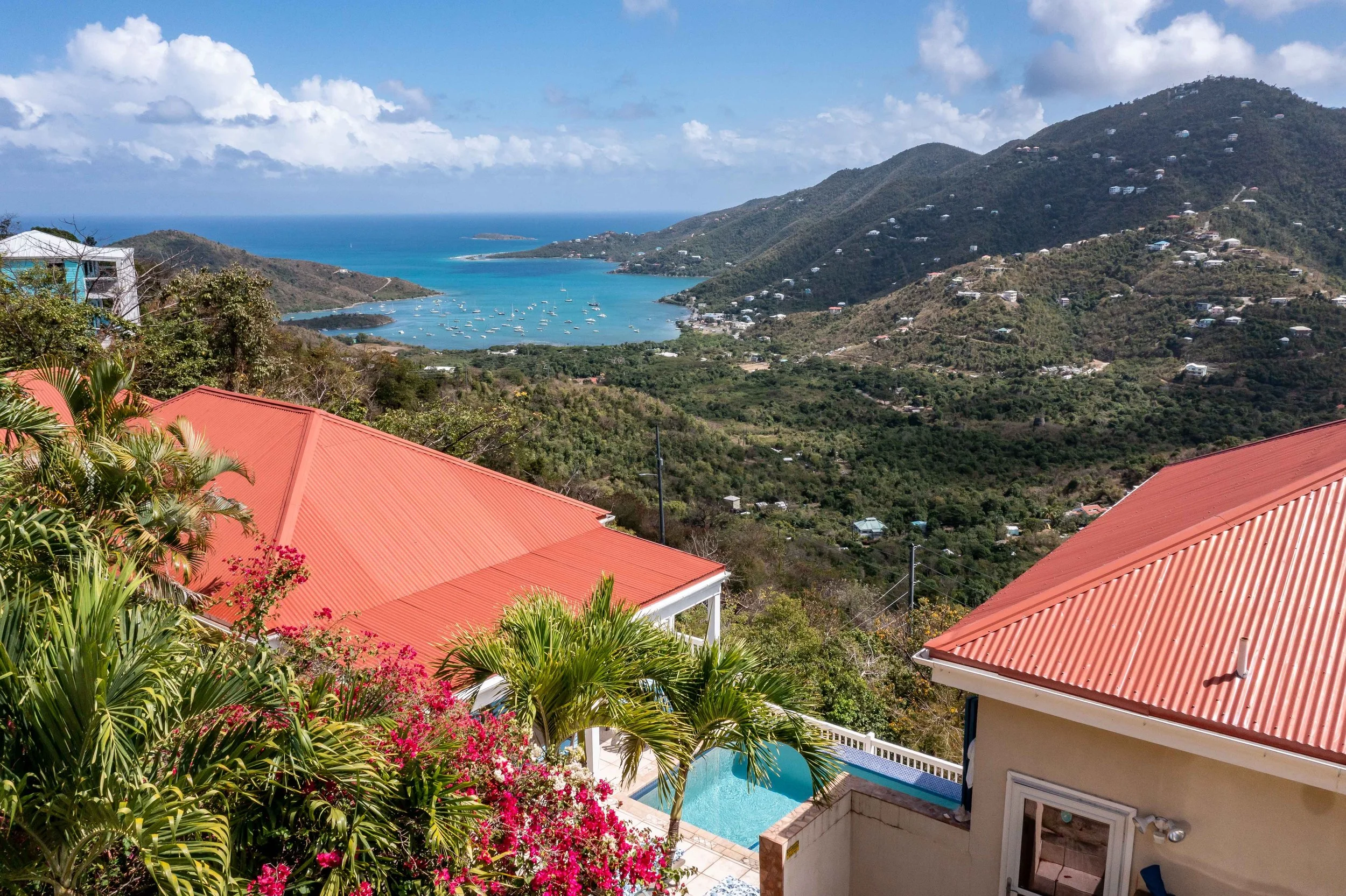 A view of a coastal landscape with hills and a bay filled with boats, seen from a house with red-tiled roofs and lush green plants.