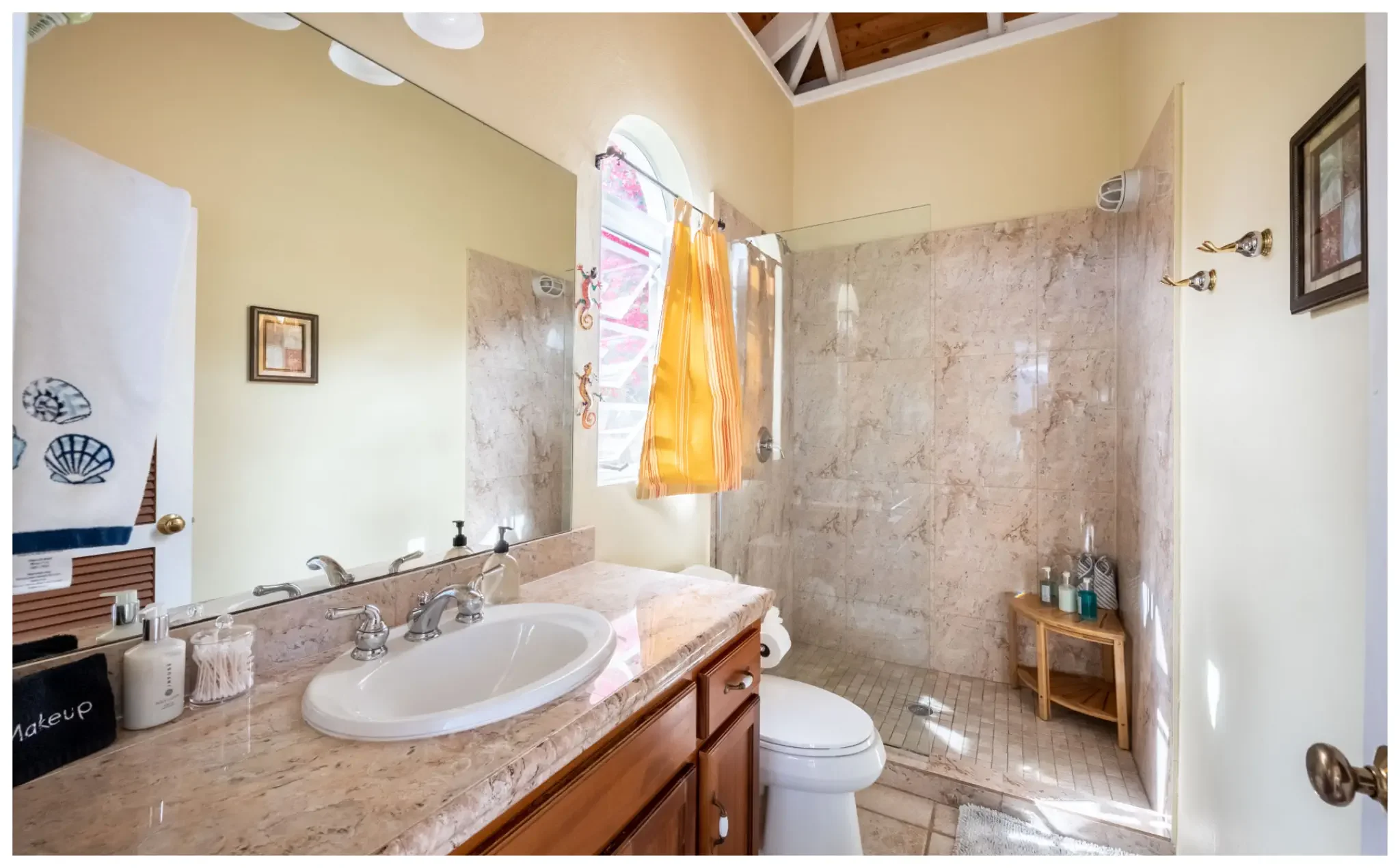 A bathroom with a marble countertop, double sinks, and a mirror. There is a window with a yellow curtain, a walk-in shower area, and various toiletries on a small wooden shelf. The walls are painted light yellow, and the ceiling has exposed wooden beams.
