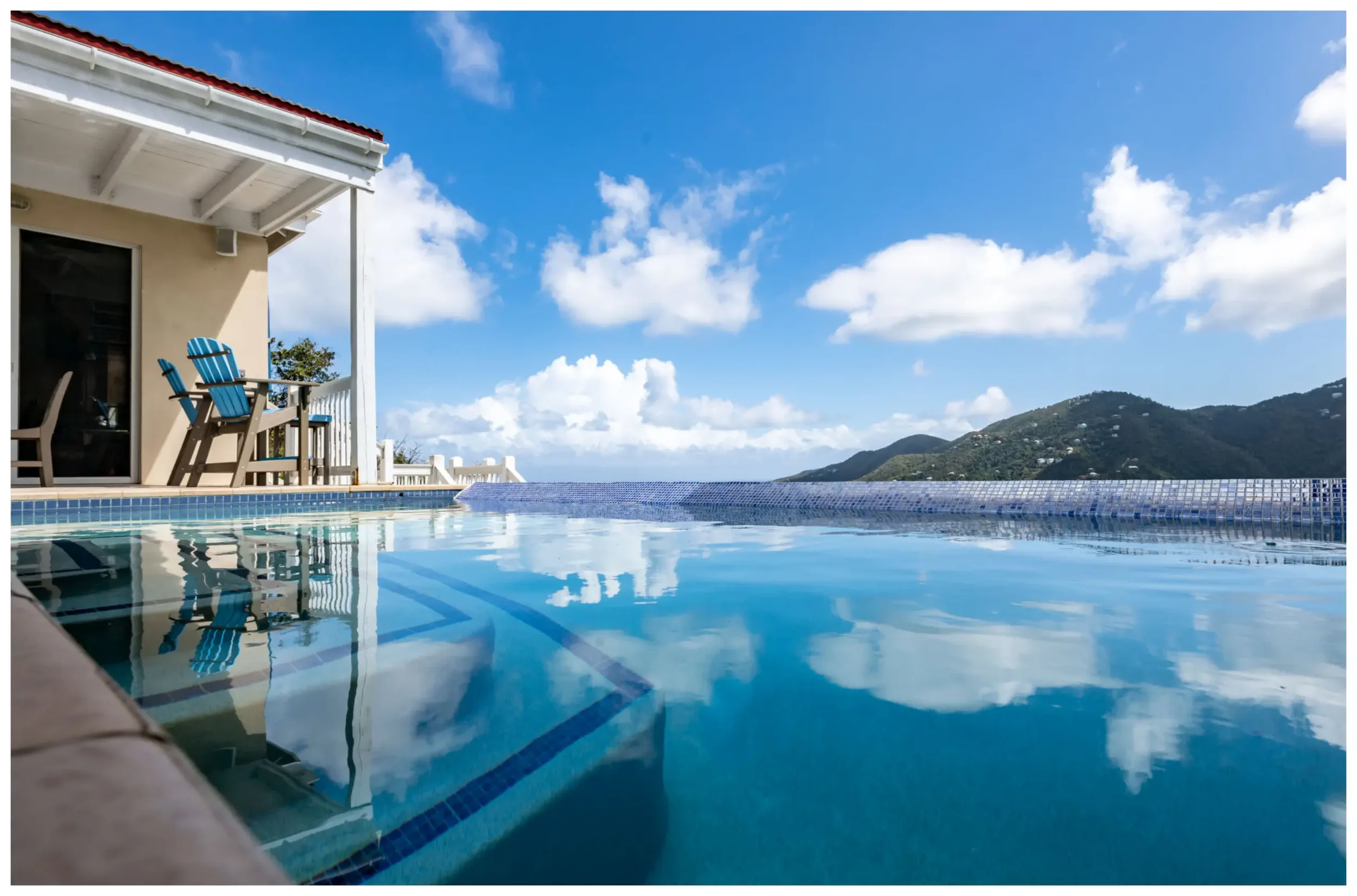 Swimming pool with mountain view and blue sky in the background, part of a house with patio chairs.