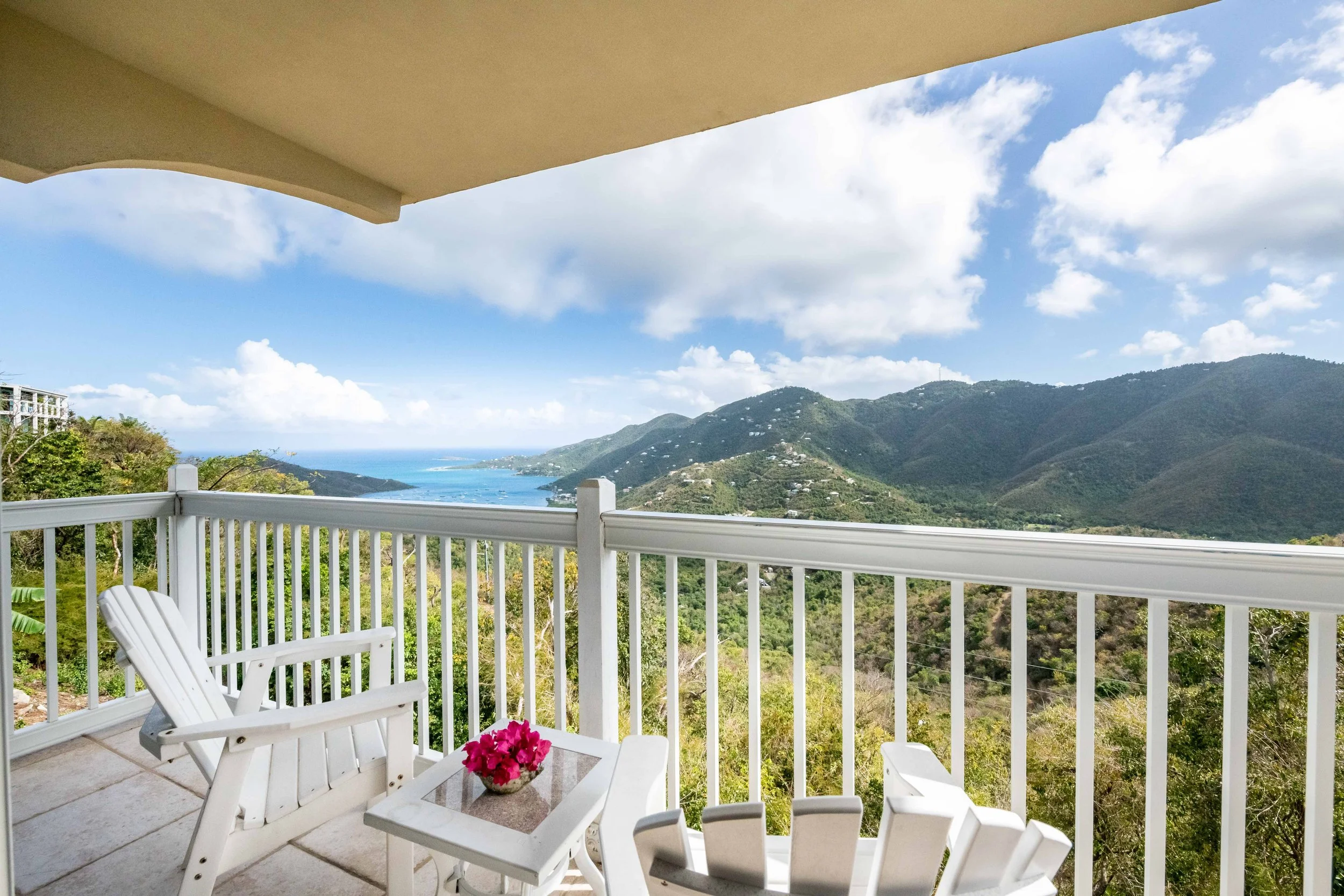 A balcony with white chairs and a table with a pink flower pot, overlooking lush green mountains and a distant ocean, under a partly cloudy sky.