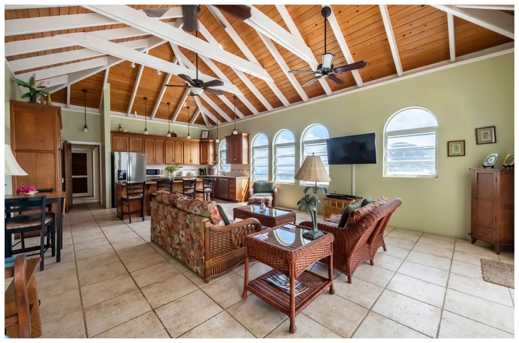 Living room and kitchen area with a vaulted wooden ceiling, ceiling fans, large arched windows, a flat-screen TV, a wicker sofa and chairs, a wooden cabinet, and various decorative items.