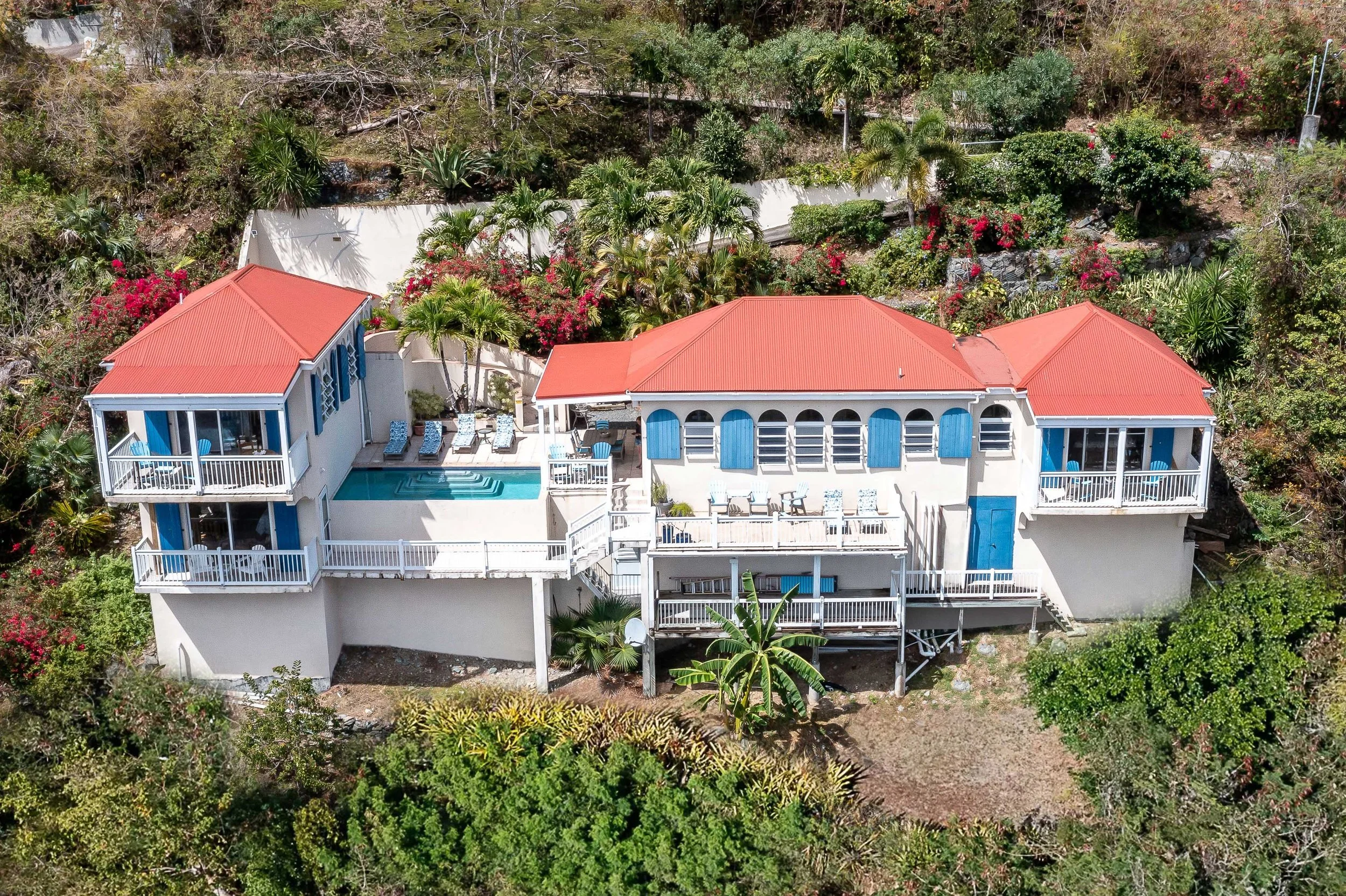 Aerial view of a white house with red roofs, surrounded by lush green trees and colorful flowering bushes. The house has multiple balconies with outdoor seating areas and a swimming pool.