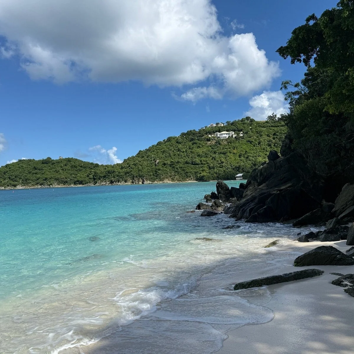 Tropical beach with white sand, turquoise water, and rocky shoreline; lush green hill with houses in the background under a partly cloudy sky.