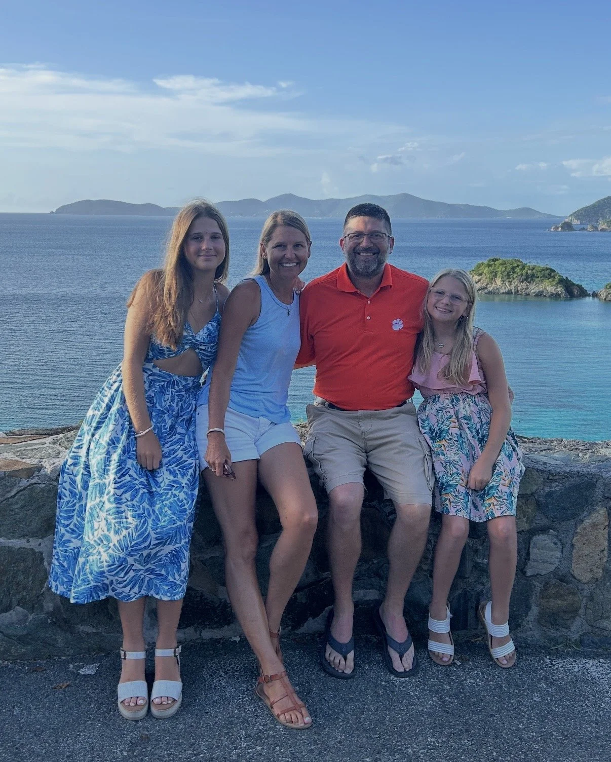 A family of four sitting on a stone wall with a scenic ocean view and islands in the background, dressed in summer clothes.