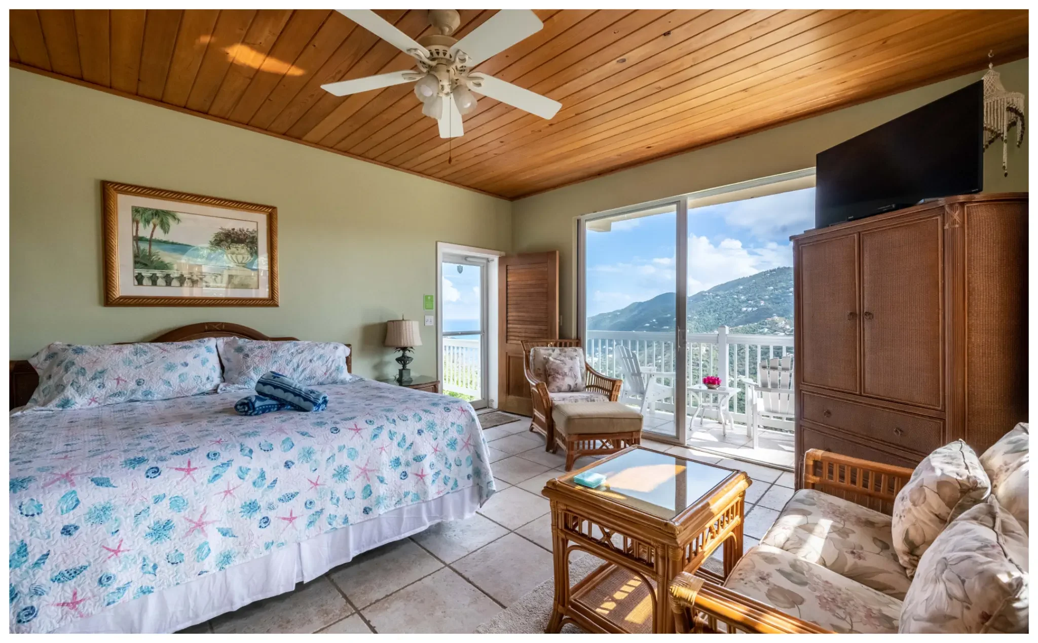 A tropical-themed bedroom with a bed covered in a seashell and starfish-patterned quilt, a wicker armchair, a small table, and large sliding glass doors opening to a balcony with ocean and mountain views, all under a wooden ceiling fan.