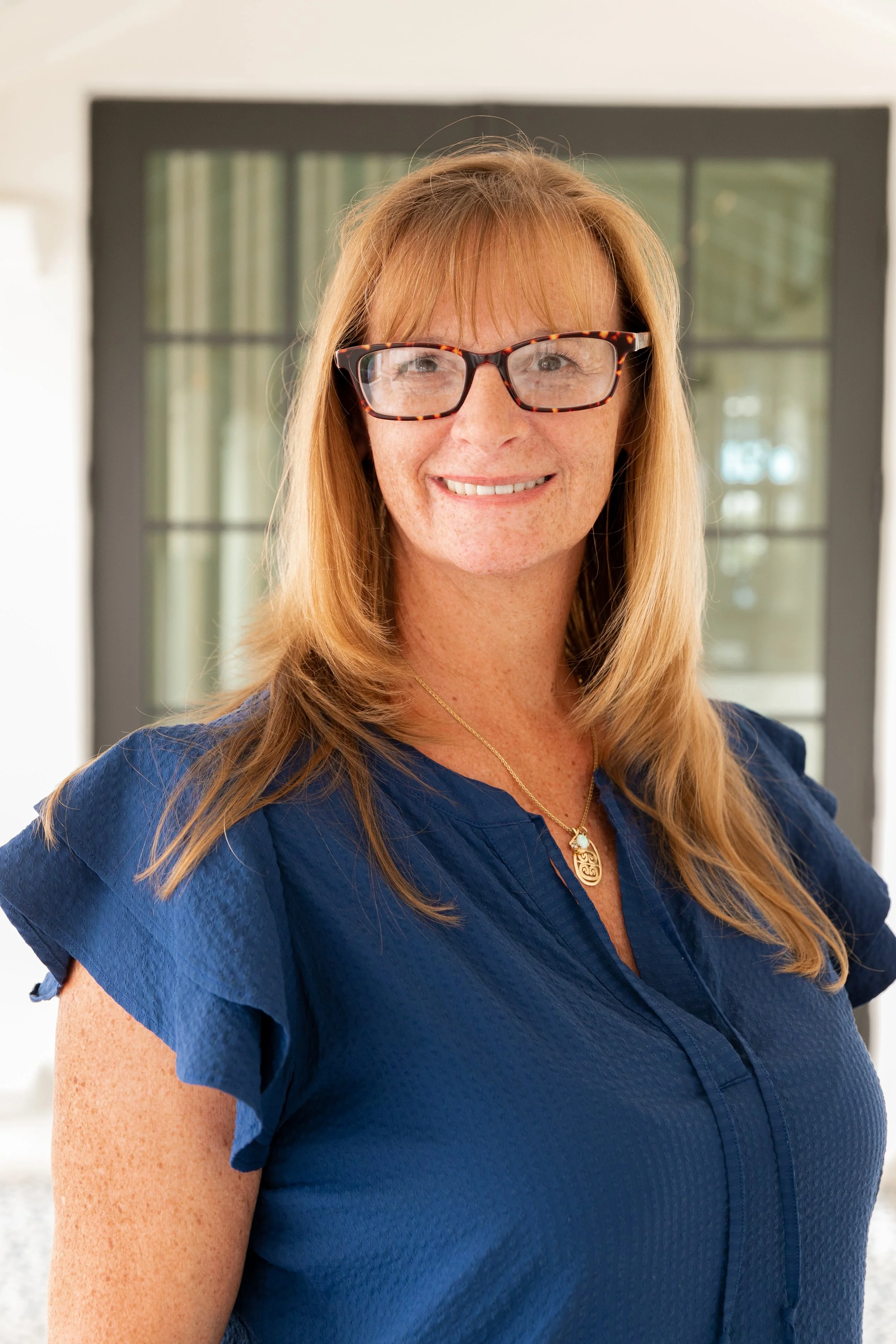 A woman with red hair, freckles, and glasses, smiling in front of a window with curtains, wearing a blue blouse and a necklace.