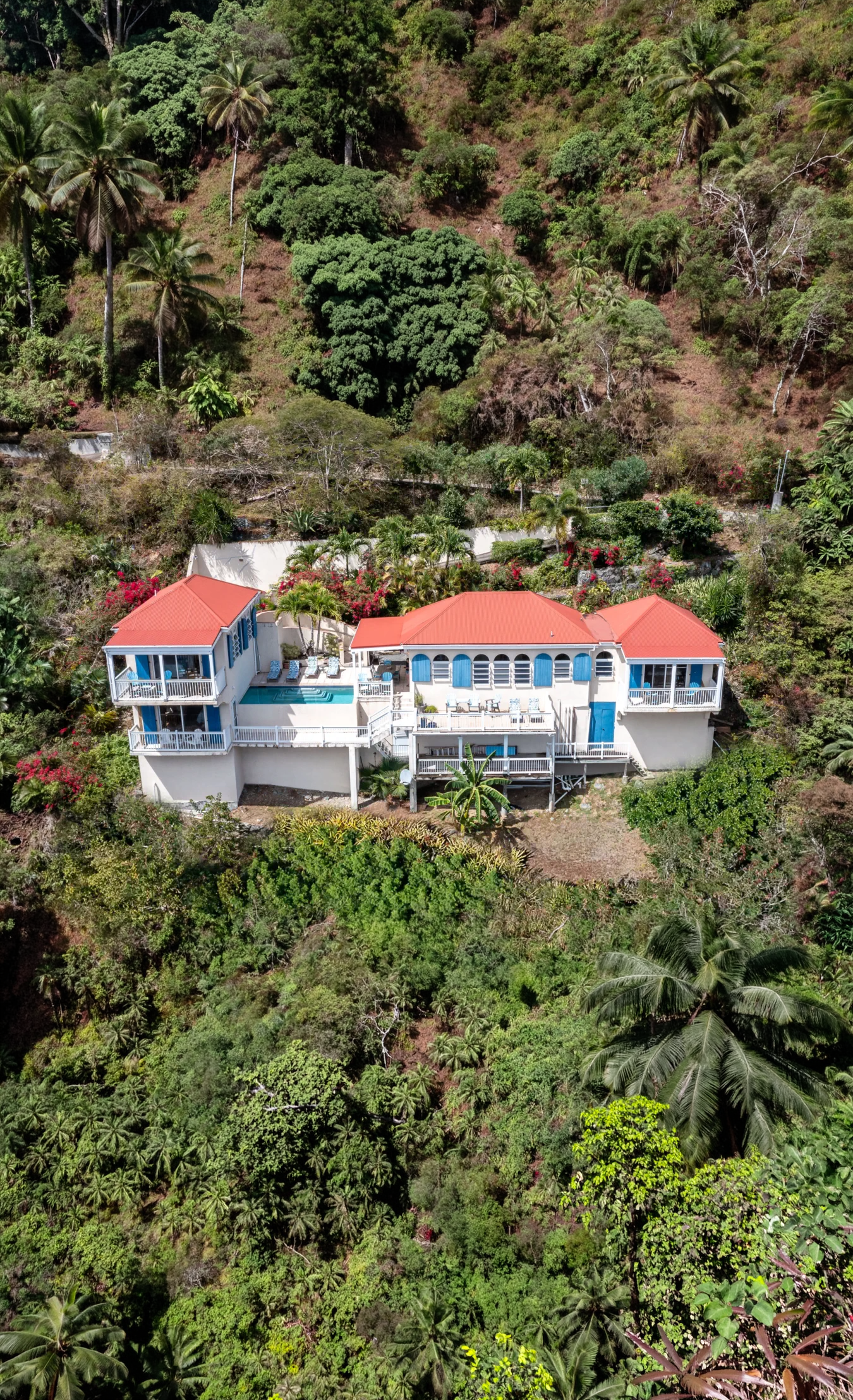 A white house with red roofs and blue shutters situated on a green hillside surrounded by lush tropical trees and vegetation.