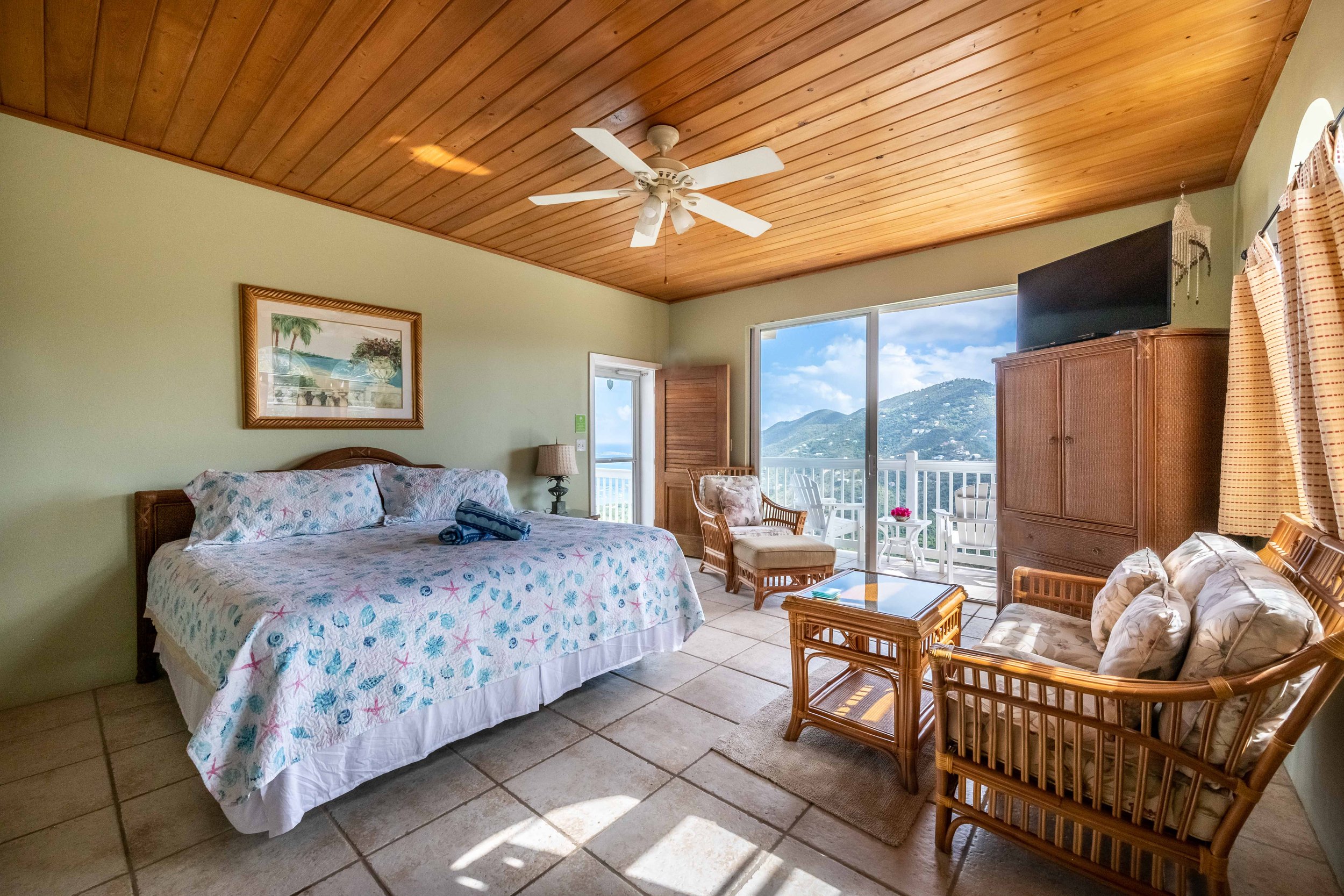 A bedroom with a tropical decor style, including a tropical-themed bedspread, wicker furniture, a ceiling fan, mountain view from the window, and artwork of palm trees.