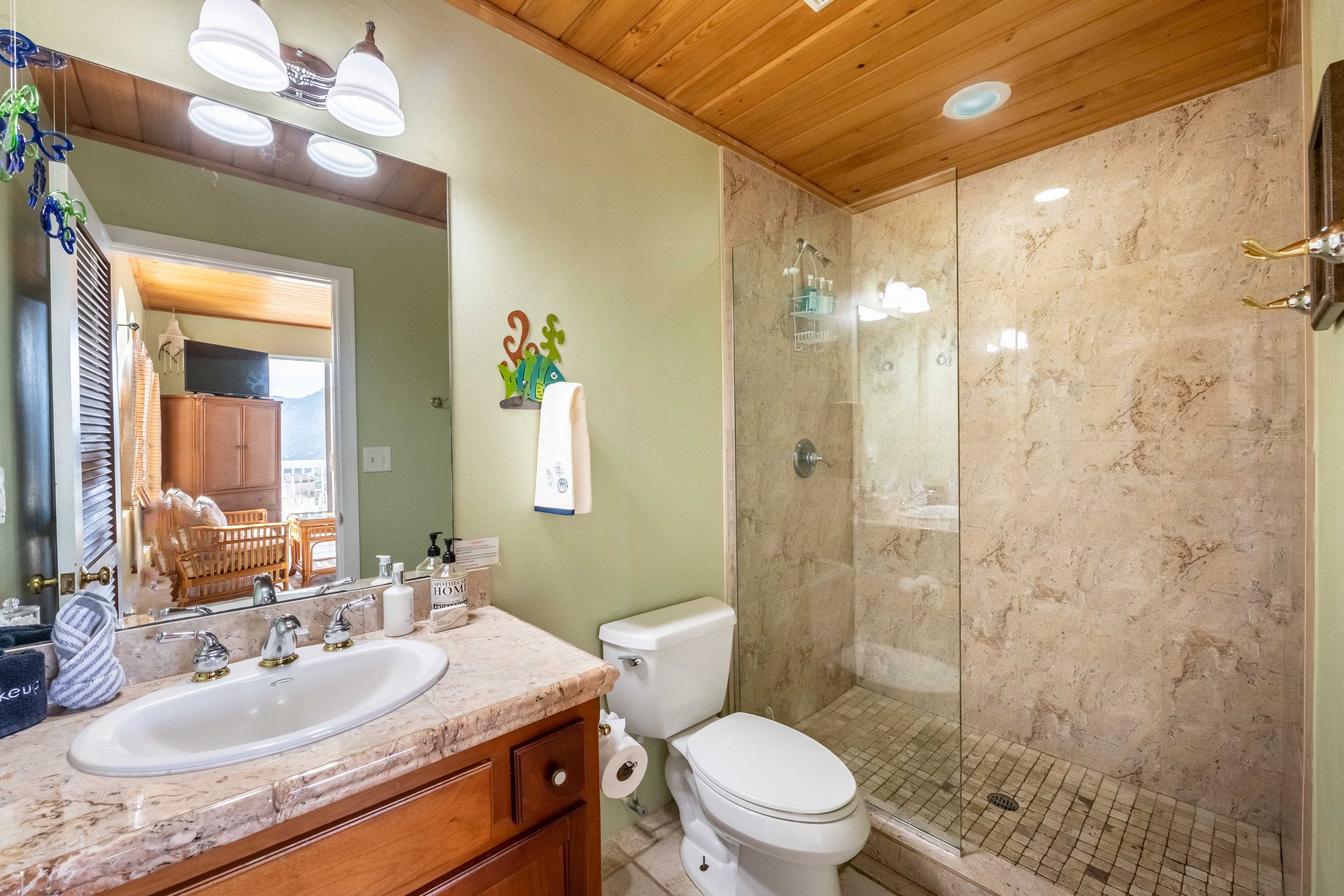 A bathroom with a double sink vanity, a mirror, a toilet, and a glass-enclosed shower. The ceiling is wood-paneled, and there is a window with wooden blinds in the adjacent room.