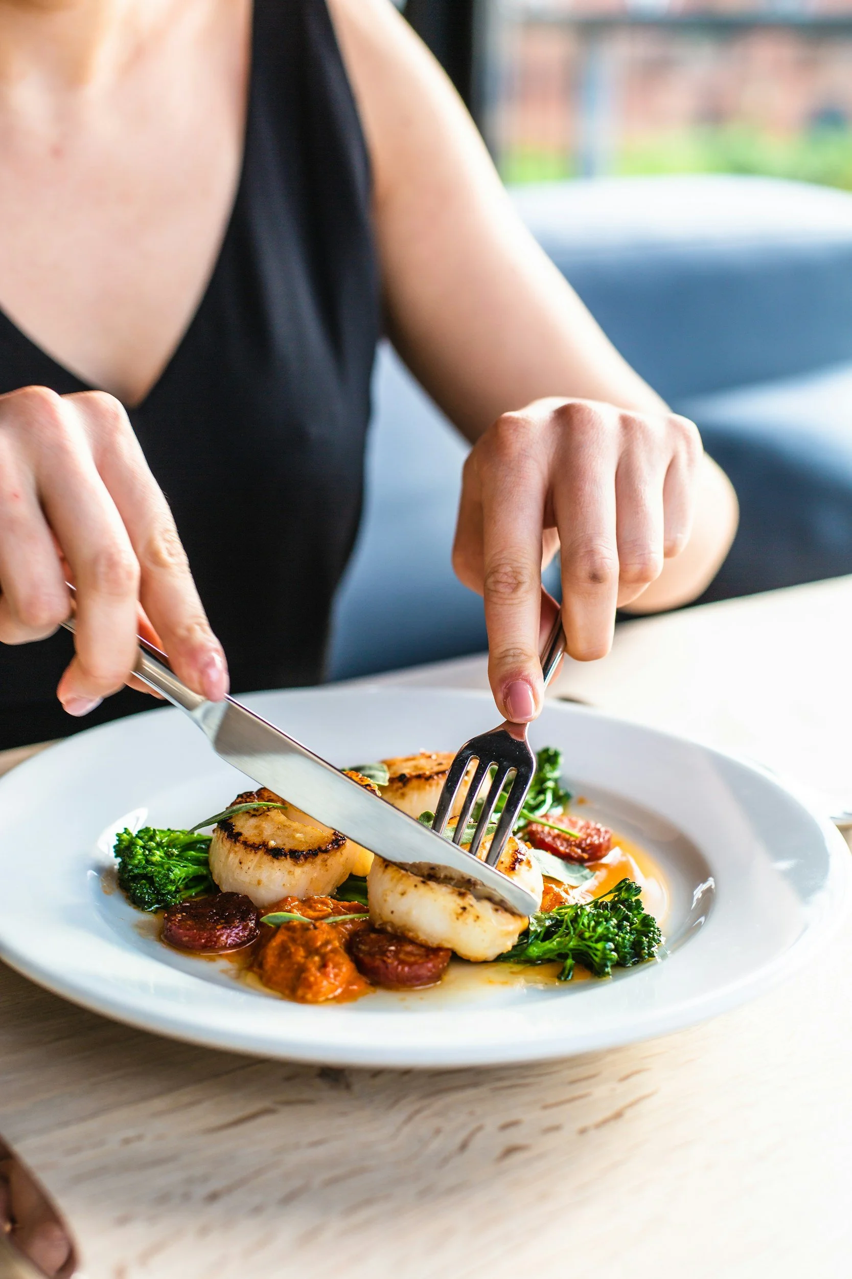 A person in a black top is eating a dish with grilled scallops, broccoli, and slices of sausage on a white plate.