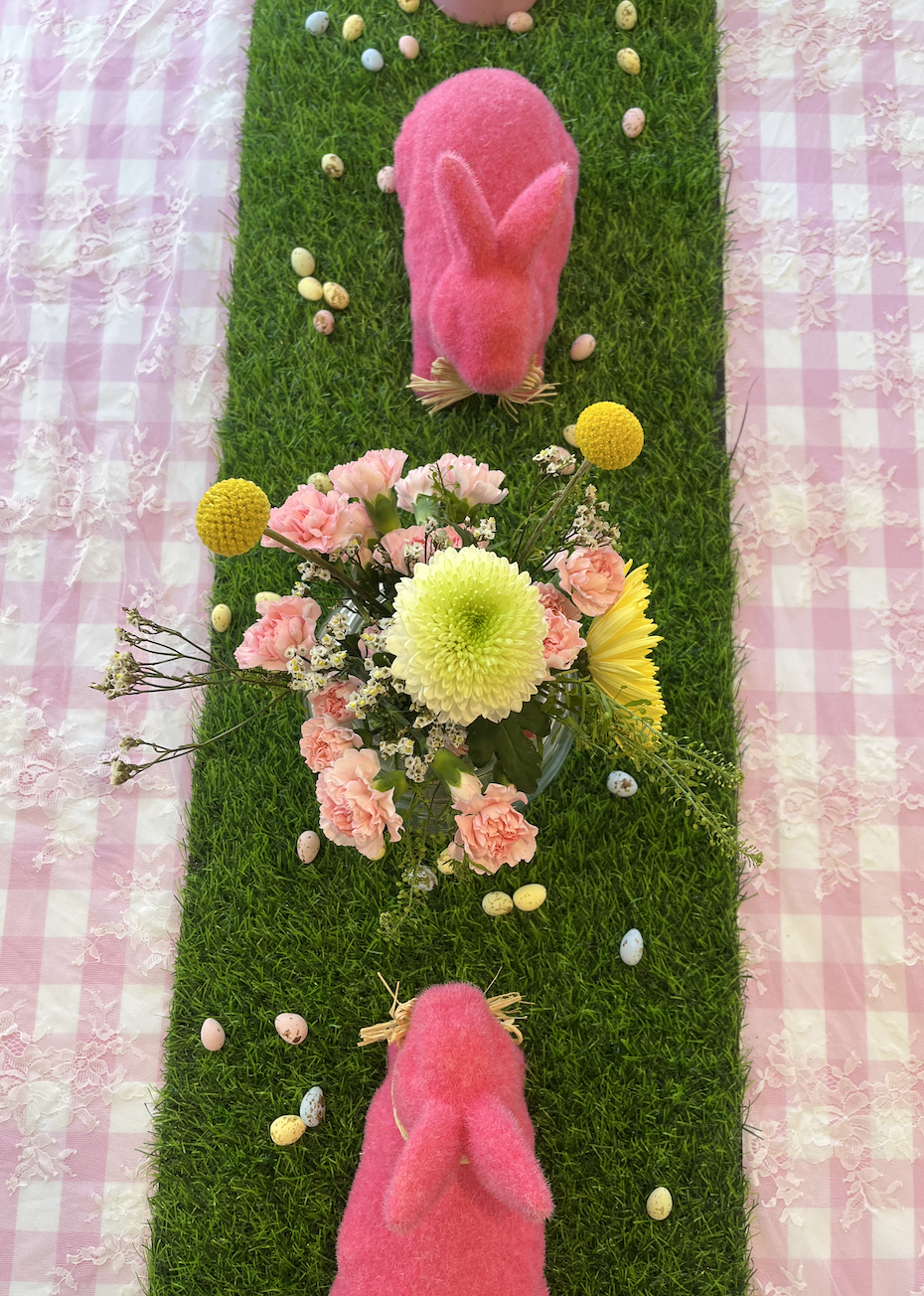 Pink plush bunny slippers on green grass with a bouquet of pink and yellow flowers and scattered pastel-colored eggs, on a pink and white checkered tablecloth.