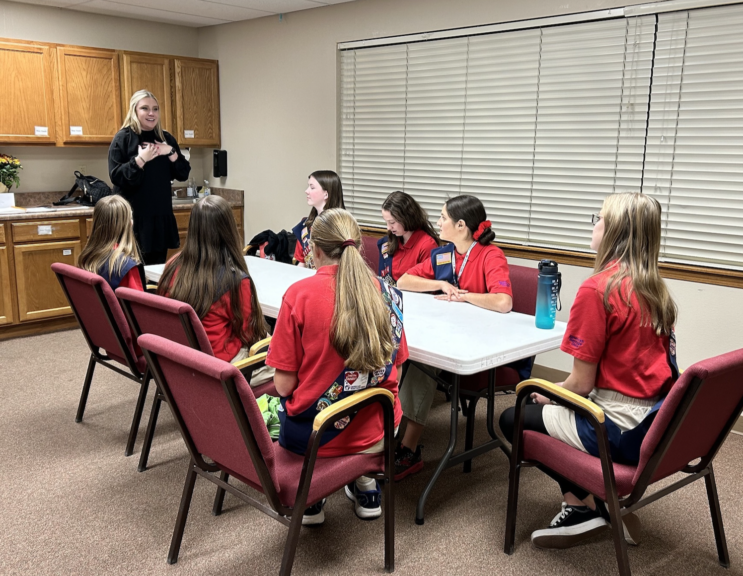 A group of young girls wearing sitting around a table in a meeting room, with one girl standing and speaking to the group.