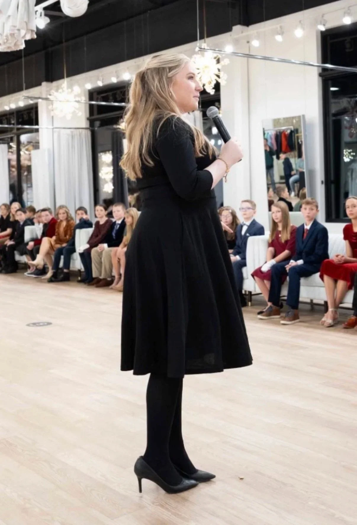 Woman in a black dress and high heels speaking into a microphone while a group of children in formal attire sit on white chairs awaiting her speech in a well-lit, modern space.