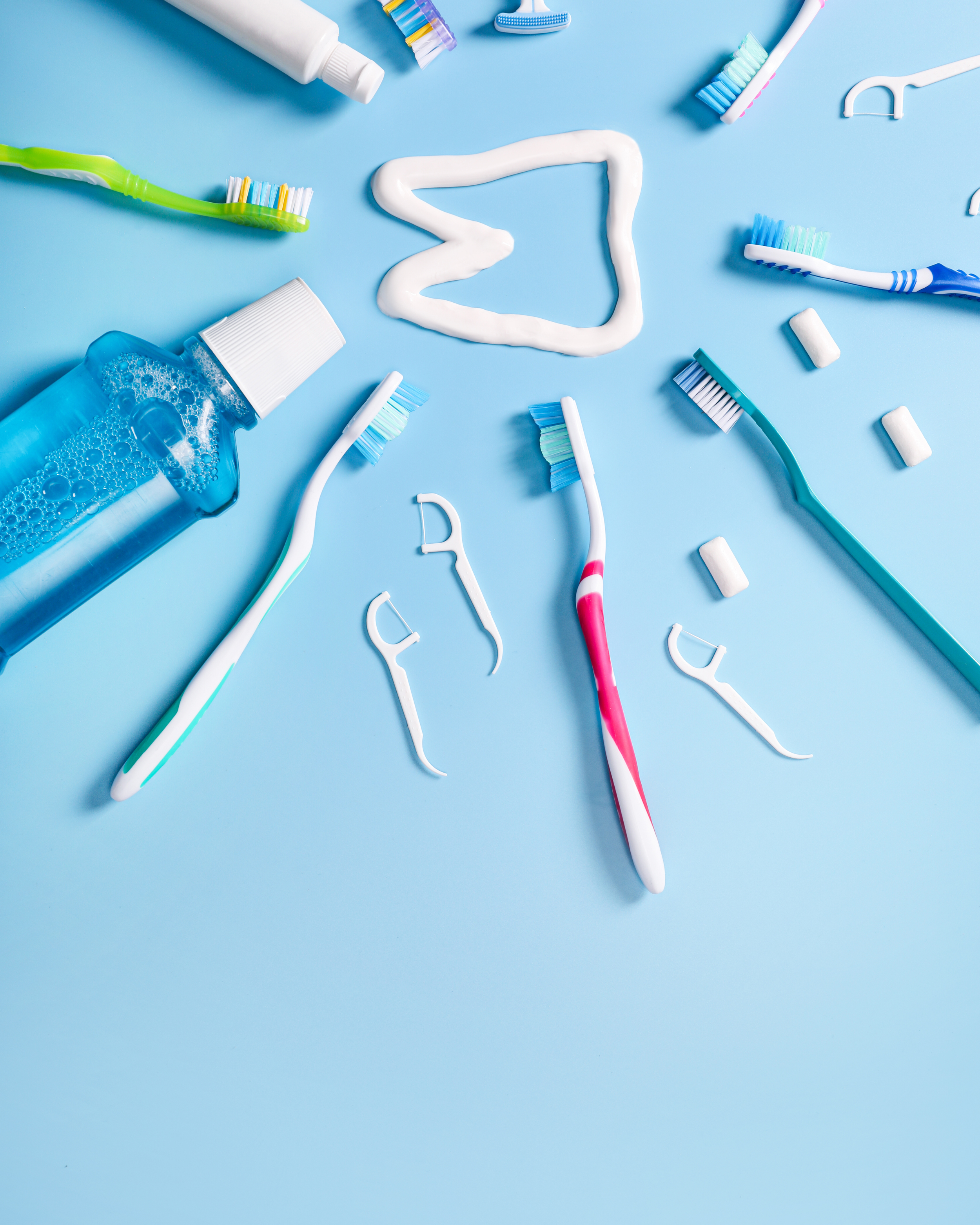Assorted toothbrushes, toothpaste, and a bottle of mouthwash arranged on a light blue surface, with a heart-shaped line of white toothpaste in the center.
