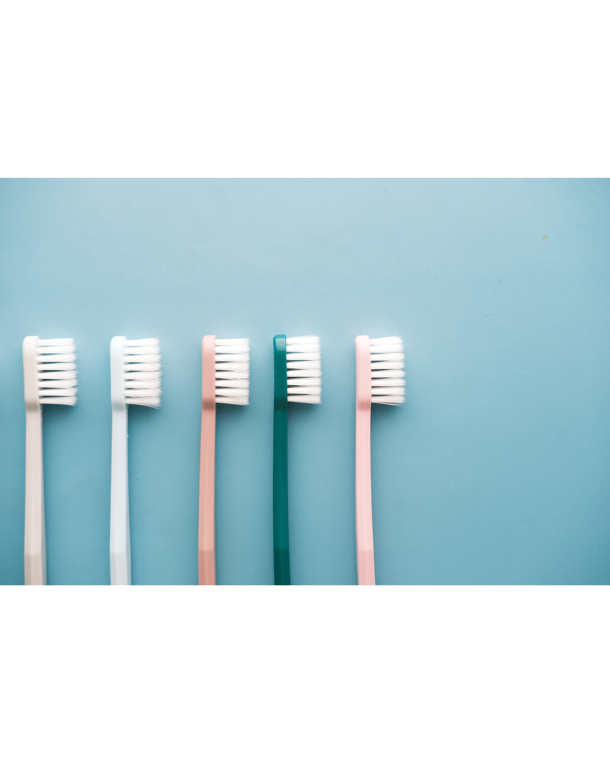 Five toothbrushes with white bristles and colored handles in pink, teal, and white, lying side by side on a light blue background.