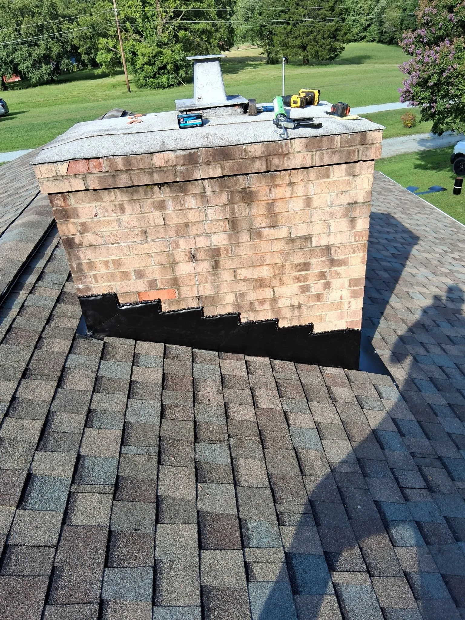 A brick chimney on a rooftop under maintenance with tools and supplies on top, including a caulking gun, batteries, and a notebook, in a residential area with grassy lawns and trees.