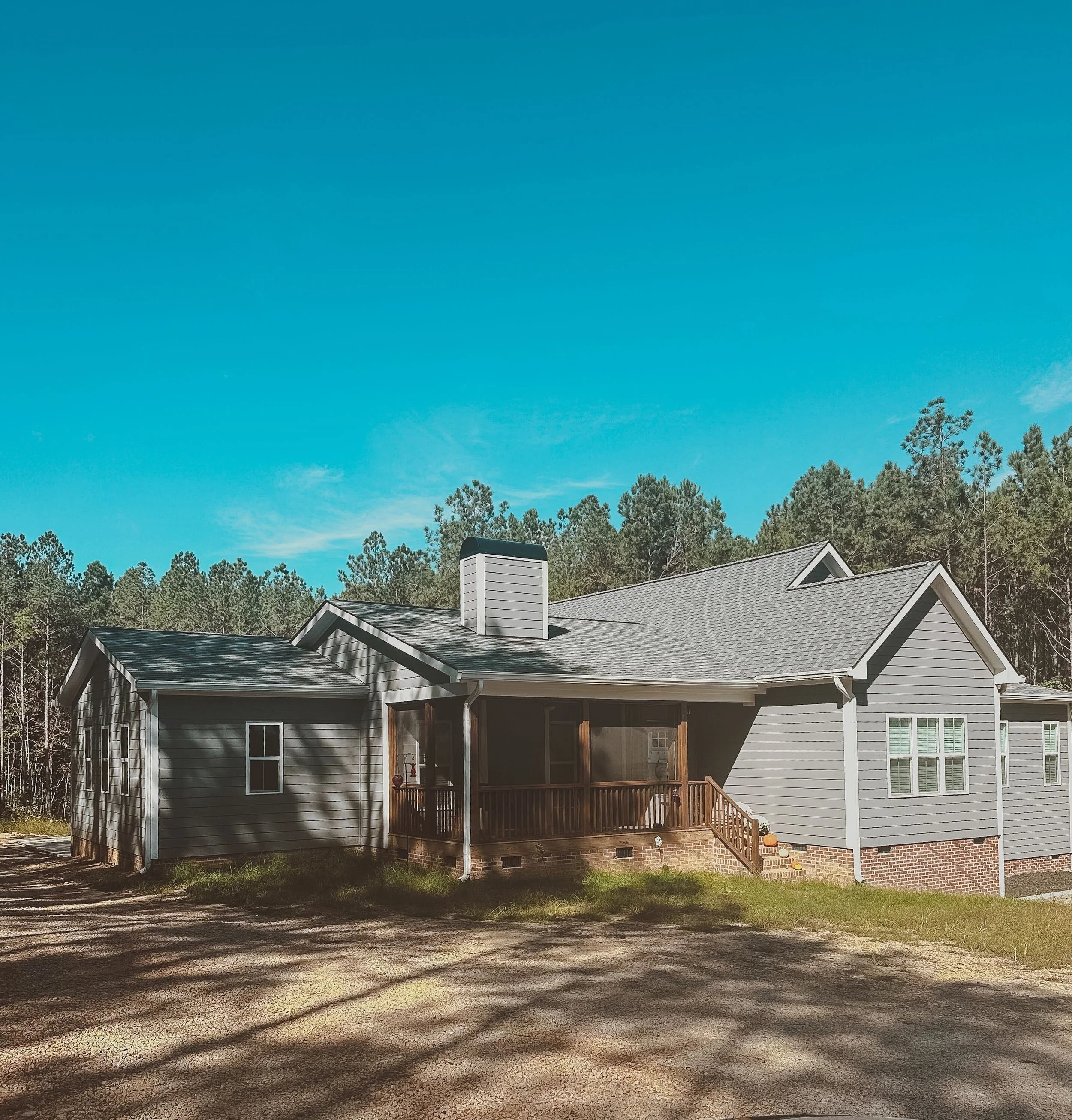 A house with a gray exterior, a screened porch, brick foundation, and a chimney, surrounded by trees under a clear blue sky.