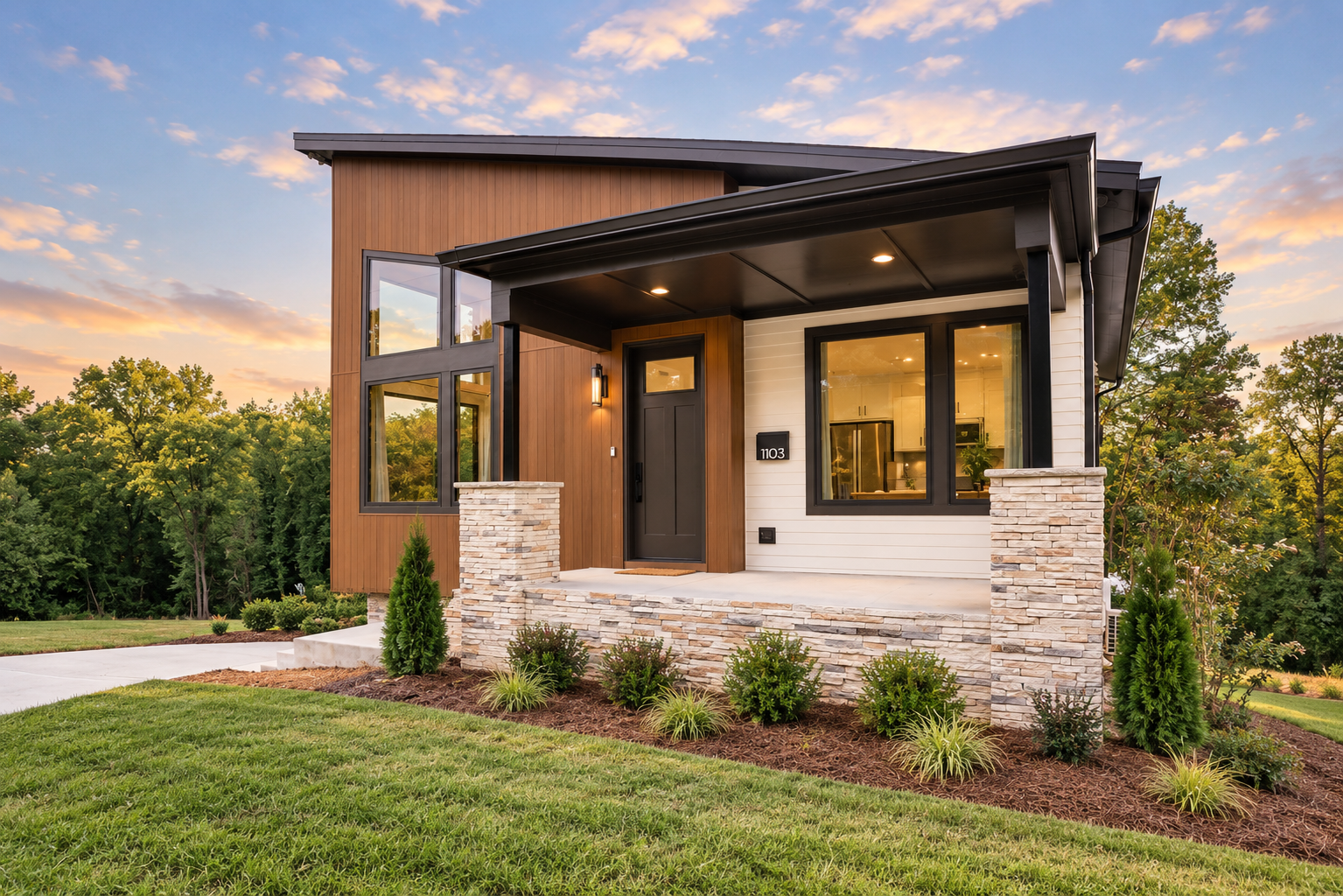 Modern house with a front porch, stone accents, and large windows, set against a background of trees and a sunset sky.
