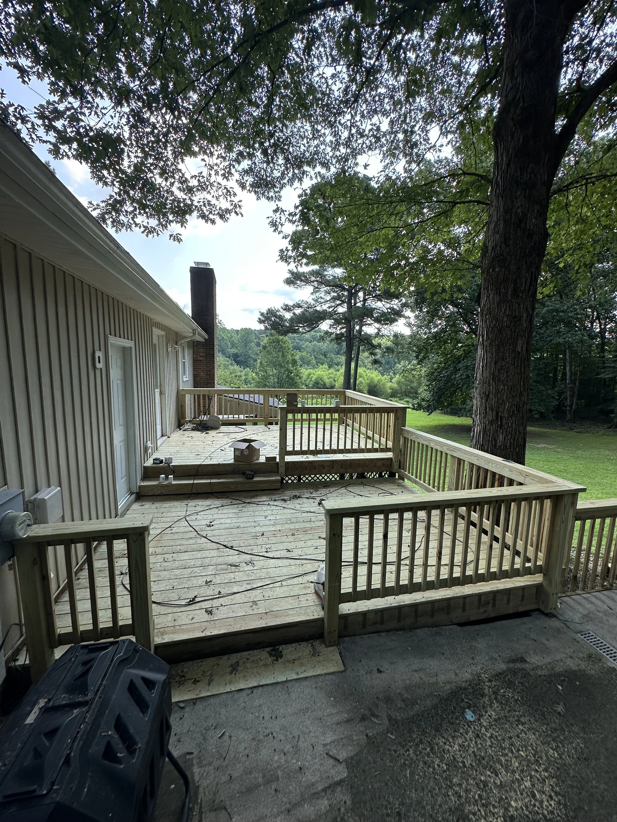 An outdoor wooden deck attached to a residential house, with surrounding trees and a grassy backyard. Construction materials and tools are visible on the deck.