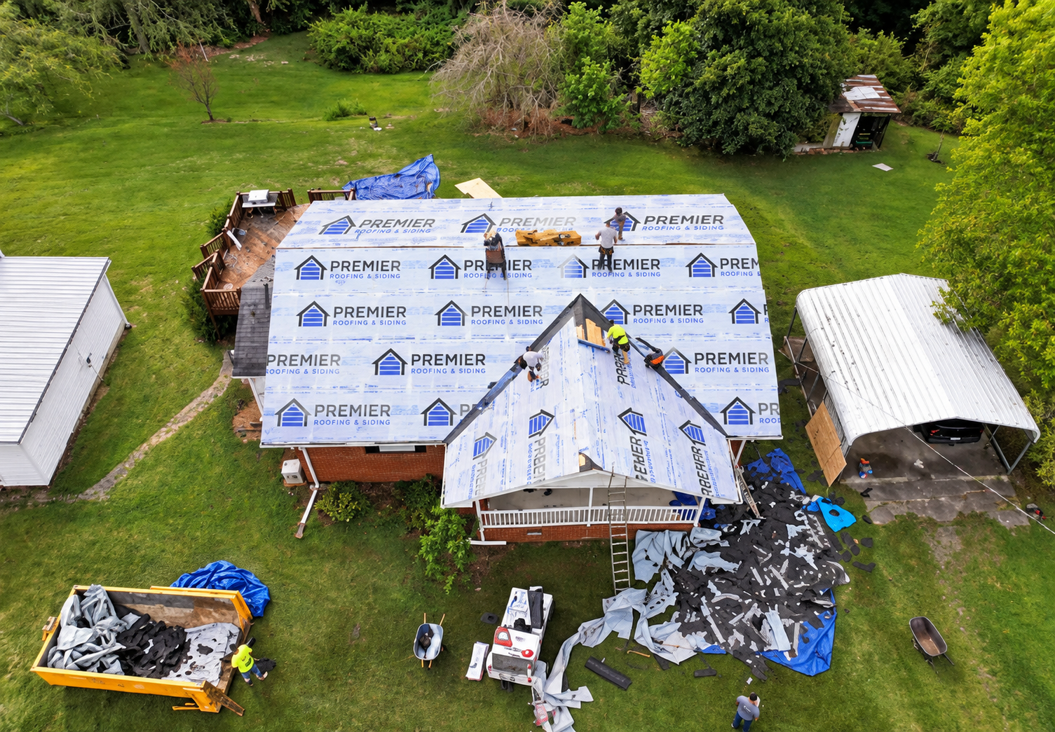 Aerial view of house roof under renovation with workers installing new roofing material. The roof is partially covered with blue underlayment labeled 'Premier Roofing & Siding.' Construction debris and equipment are scattered around the yard, with a covered parking area to the right and garden area to the left.