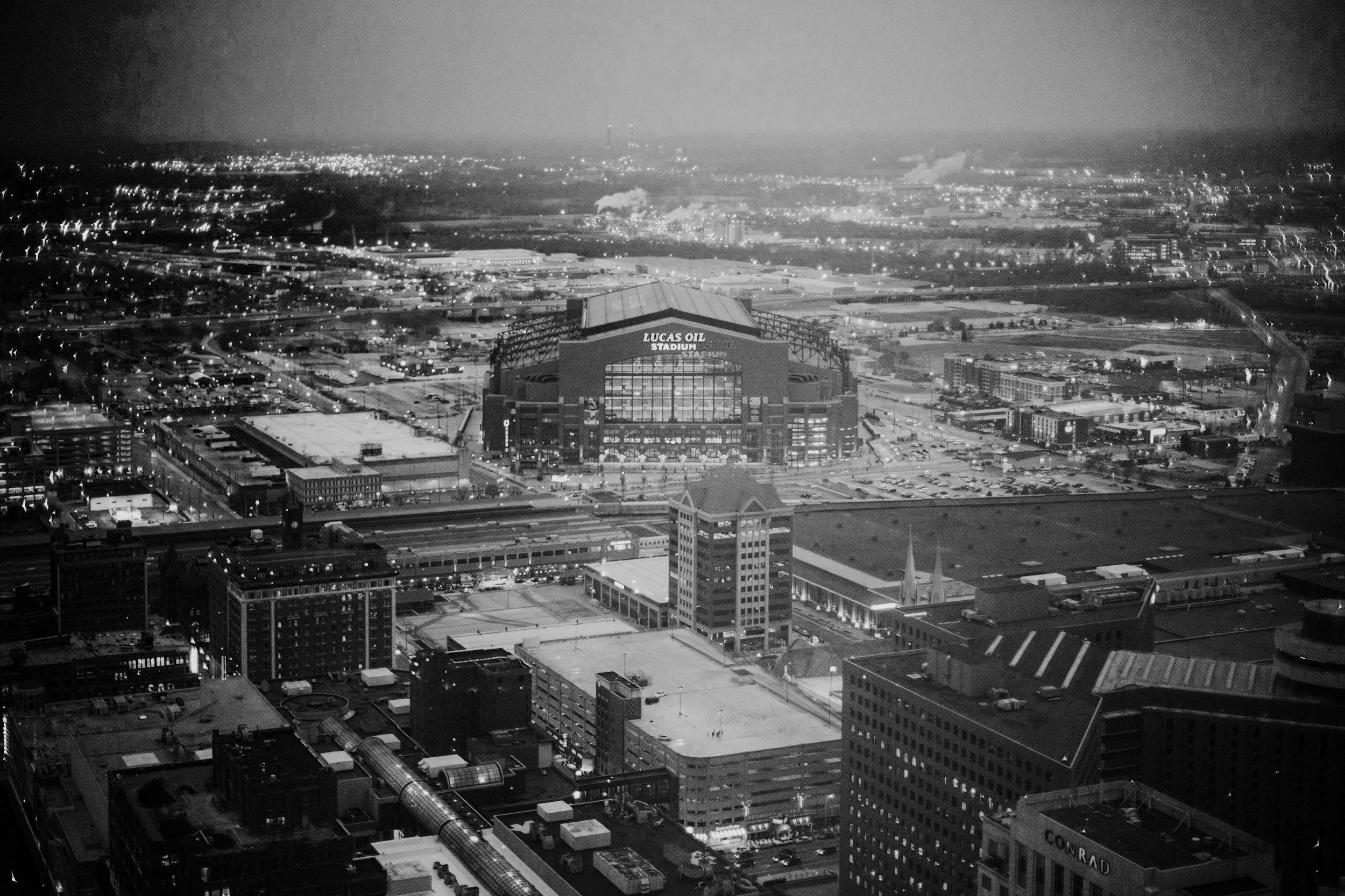 Nighttime aerial view of a cityscape featuring a stadium labeled 'Lucas Oil Stadium', surrounded by buildings, roads, and city lights in the distance, with a hazy sky.