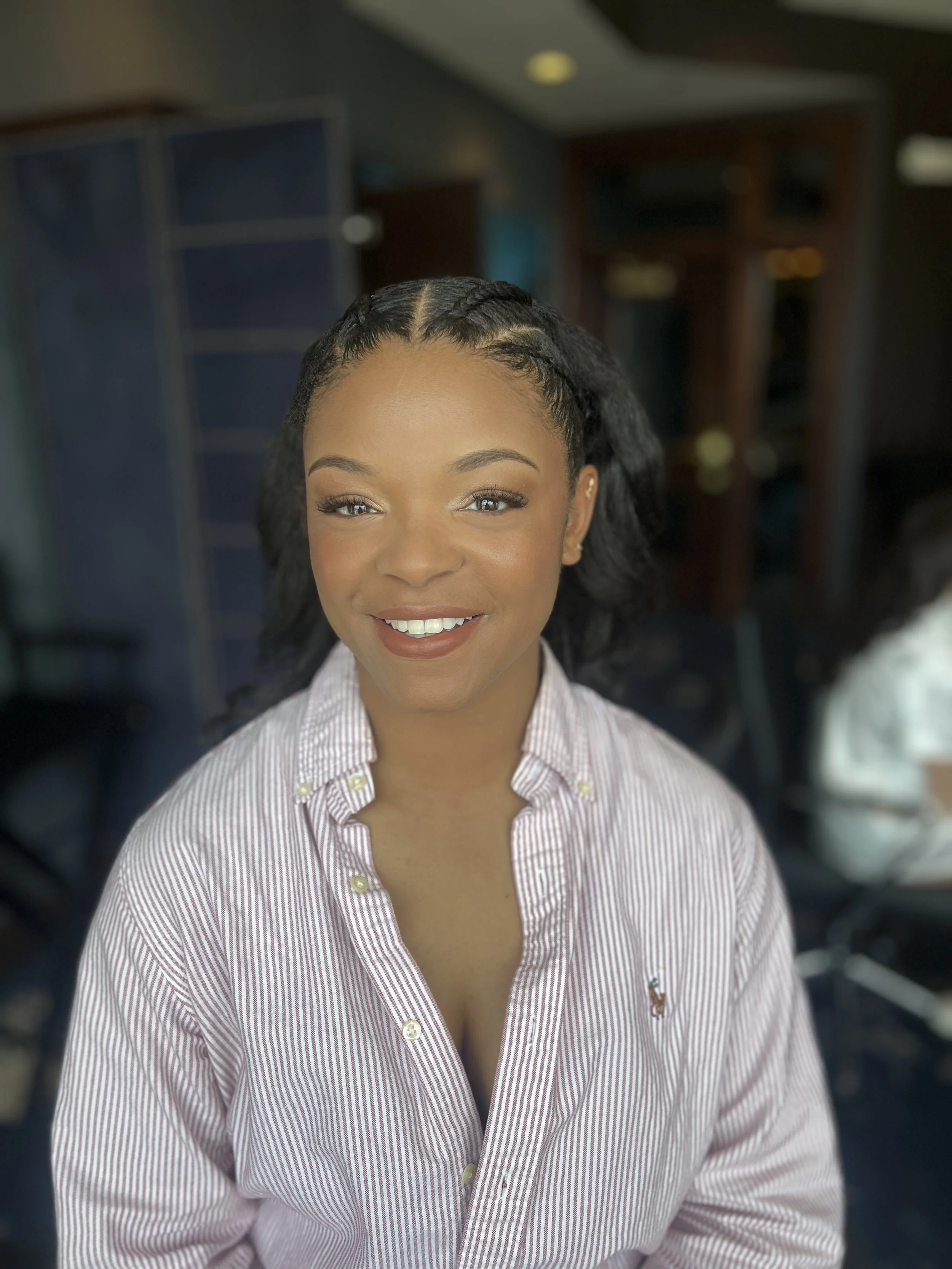 A young woman smiling, wearing a striped button-up shirt, with dark braided hair, sitting in a modern indoor setting with blurred furniture in the background.