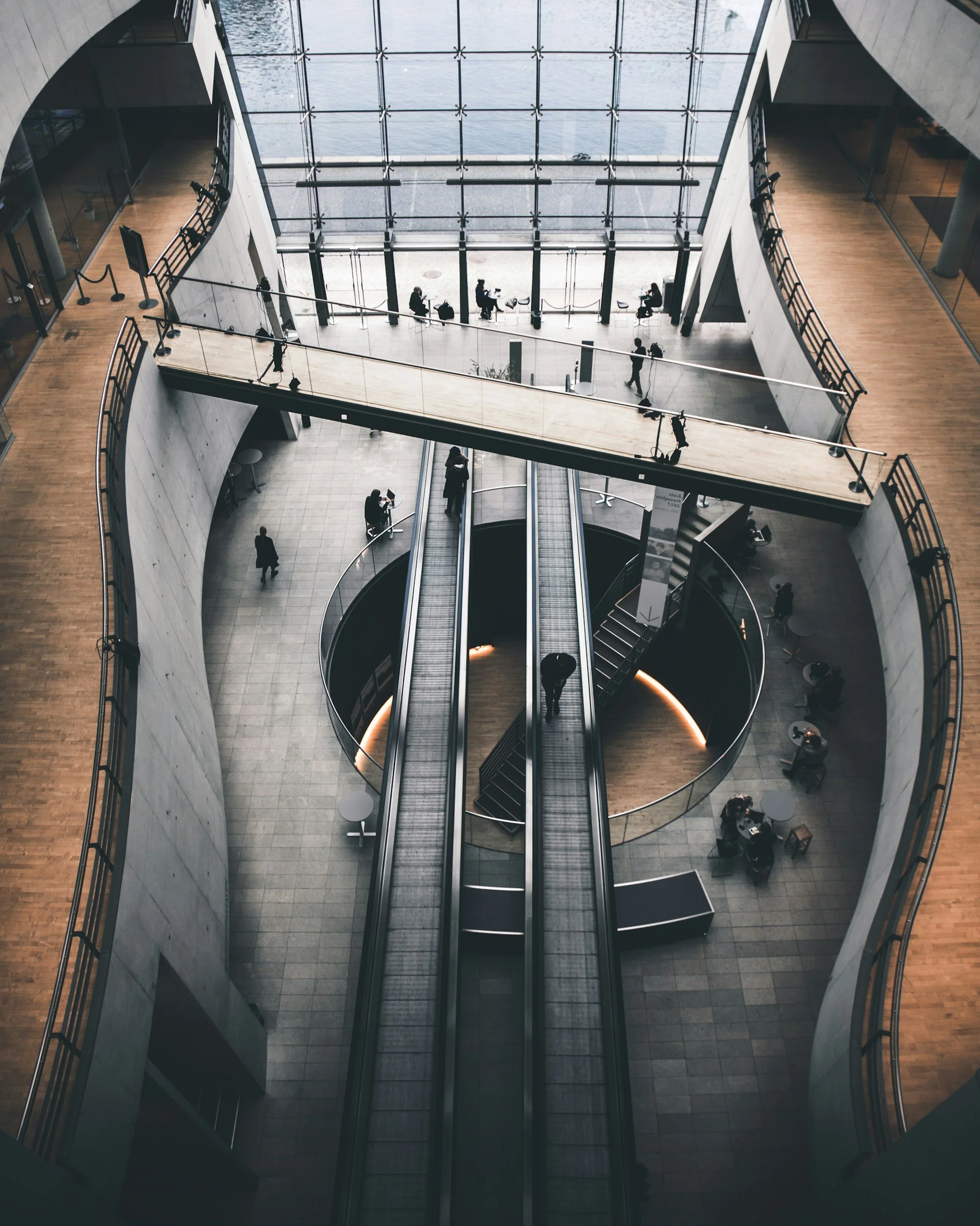 Interior of a modern building seen from an upper floor, featuring a large glass window, multiple people walking and sitting, and an escalator in the center.