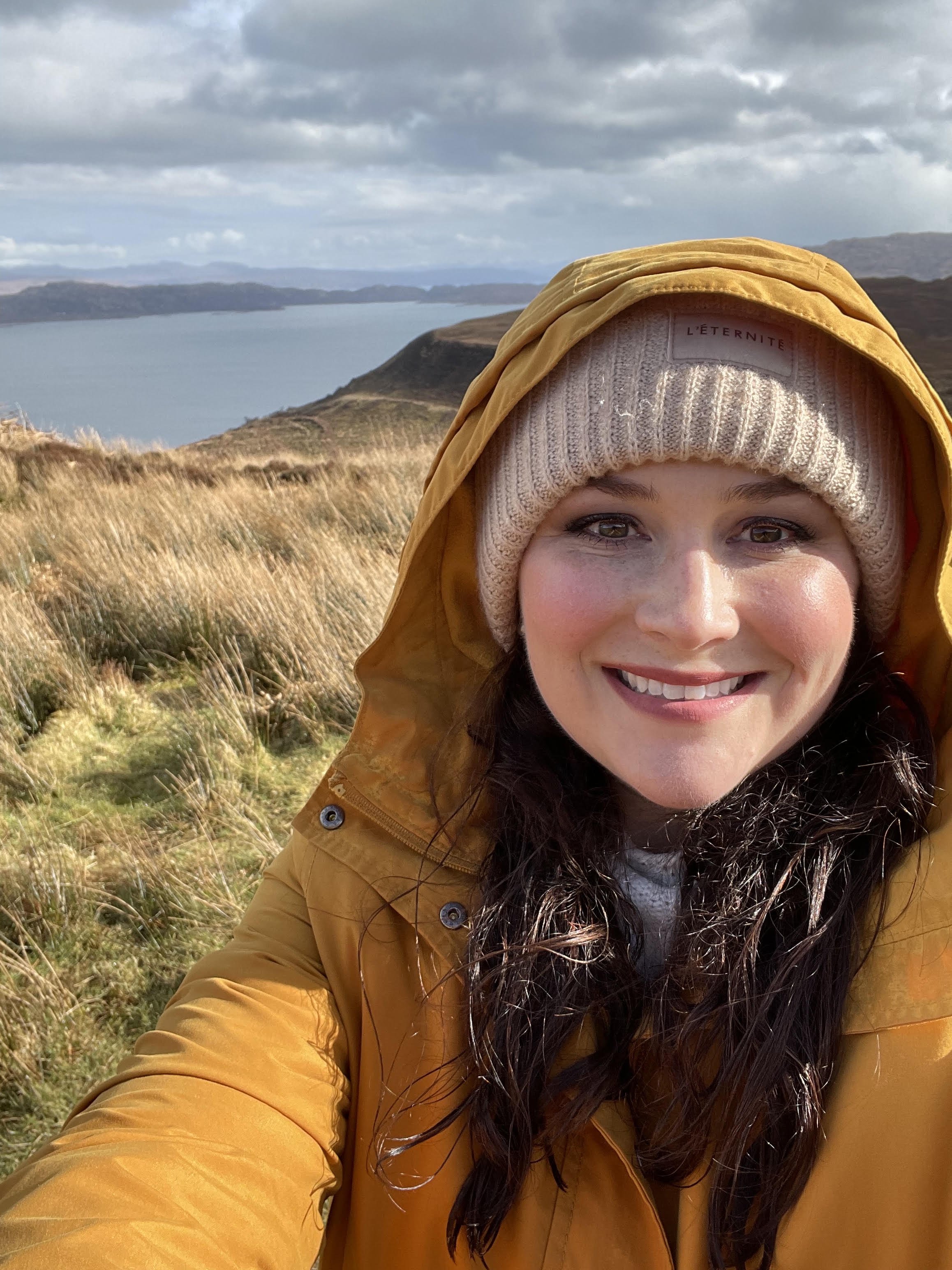 Close-up of a smiling woman with dark hair, wearing a beige knit hat and a yellow jacket, outdoors in a grassy landscape with a lake and hills in the background.