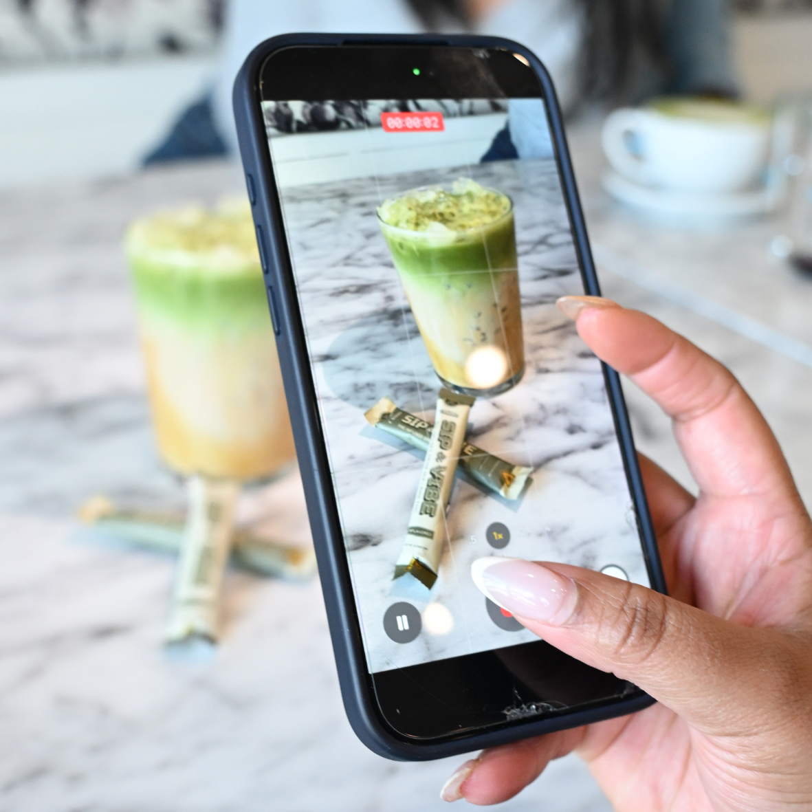 A person is taking a photo of a layered green and yellow drink on a marble table with a smartphone, with snacks and a coffee cup in the background.