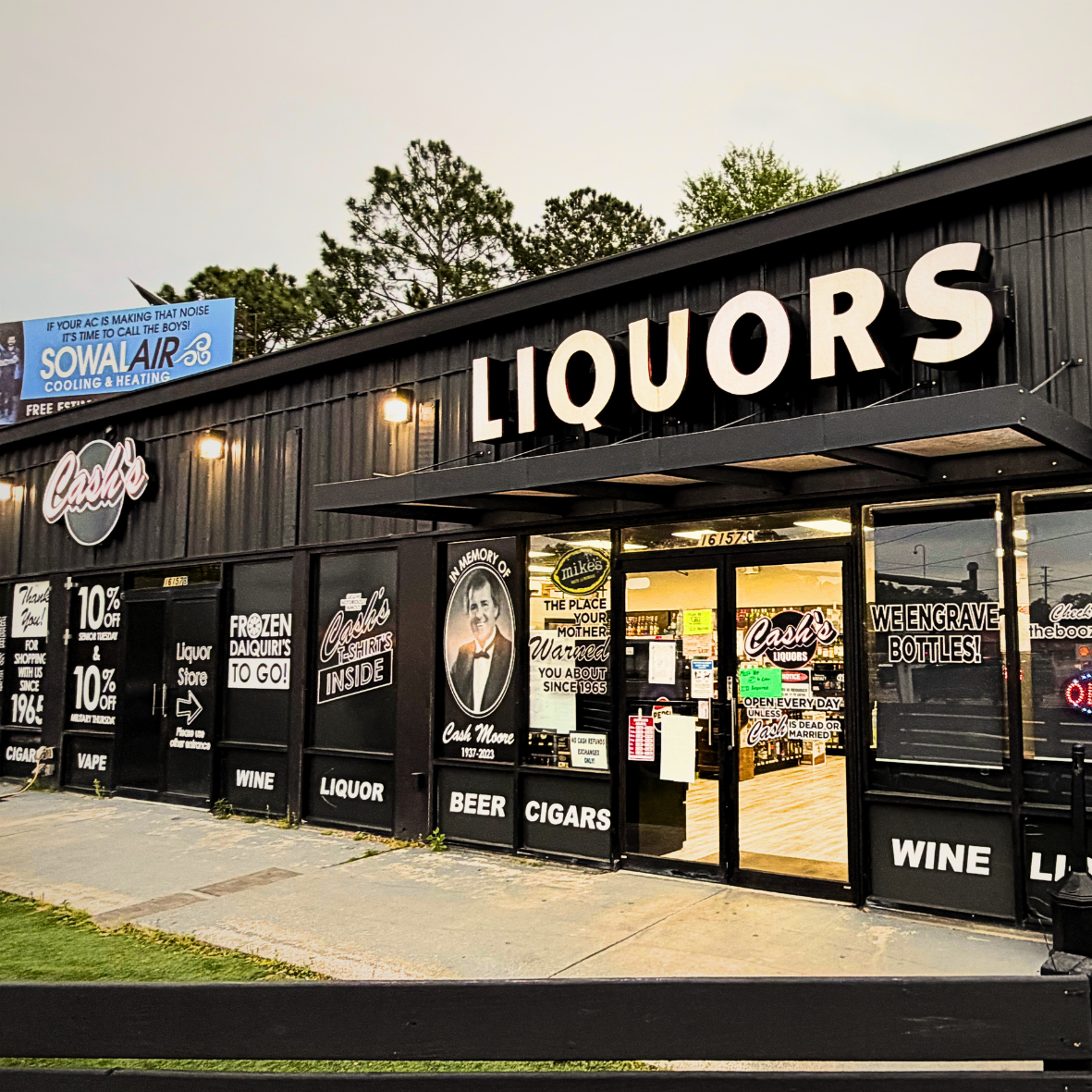 Exterior of a liquor store with large 'LIQUORS' sign on black building. Windows display signs for wine, beer, cigars, and a portrait of Cash Moye, with promotional signage.