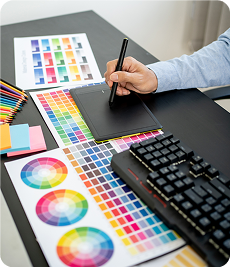 A person using a stylus on a graphics tablet surrounded by color swatches, charts, and a keyboard on a desk.